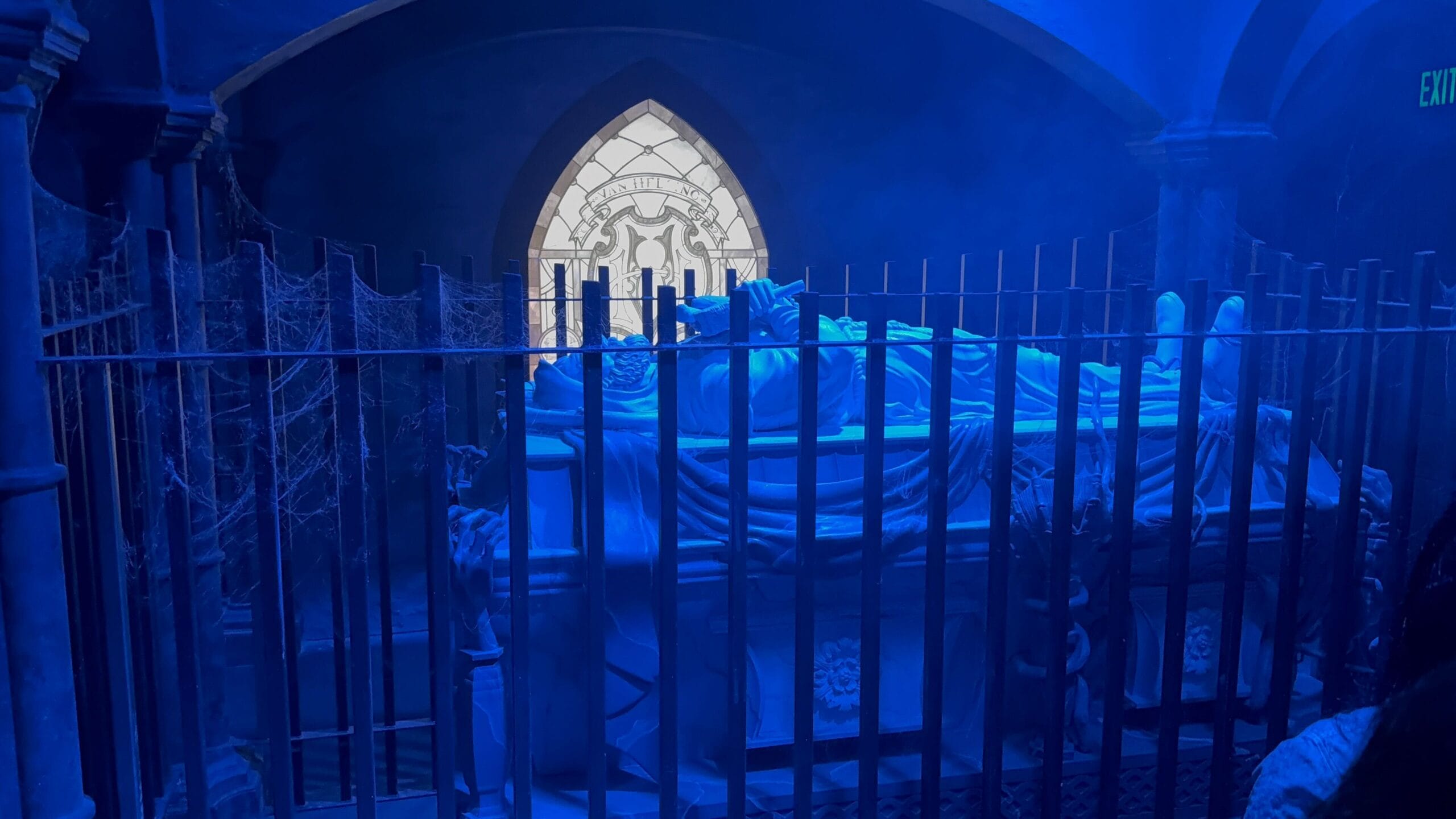 A statue lies on a tomb behind iron bars, bathed in blue light—like a scene from Universal Horror Unleashed.
