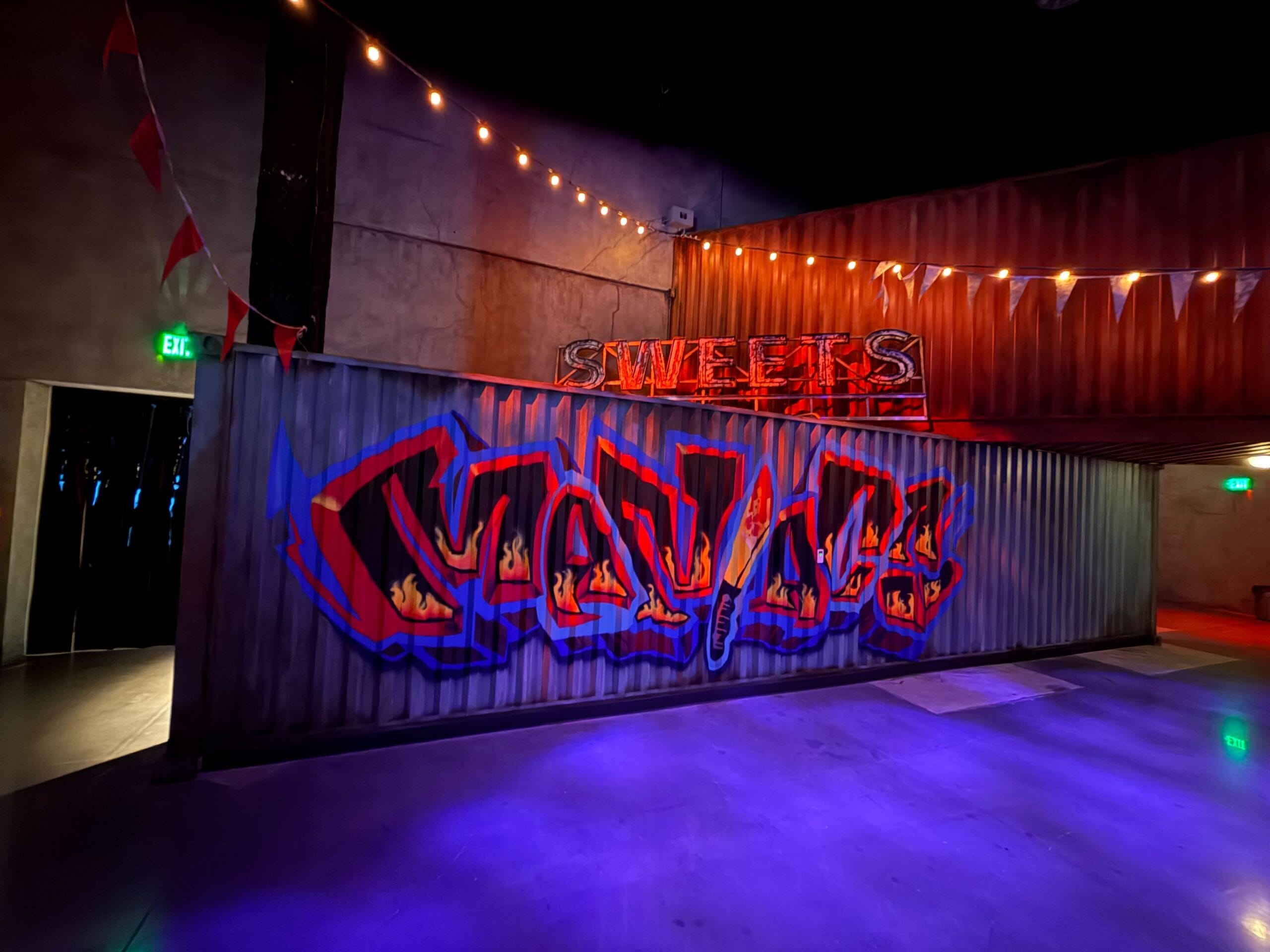 Graffiti art on a corrugated wall under string lights, with a neon "SWEETS" sign in a dimly lit Las Vegas indoor space.