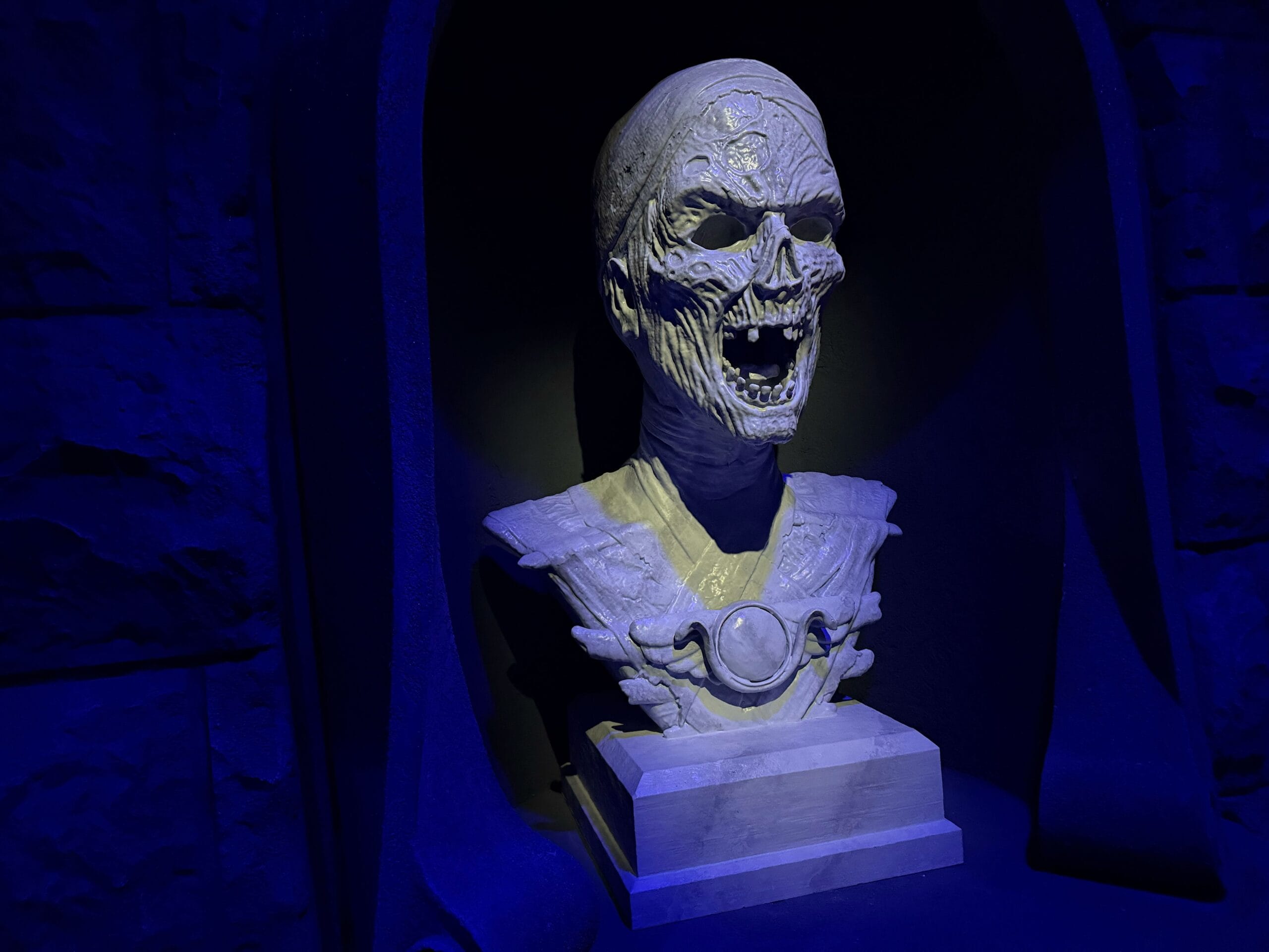 A ghoulish, skeletal bust lit by eerie blue light evokes Universal Horror Unleashed against a dark stone background.