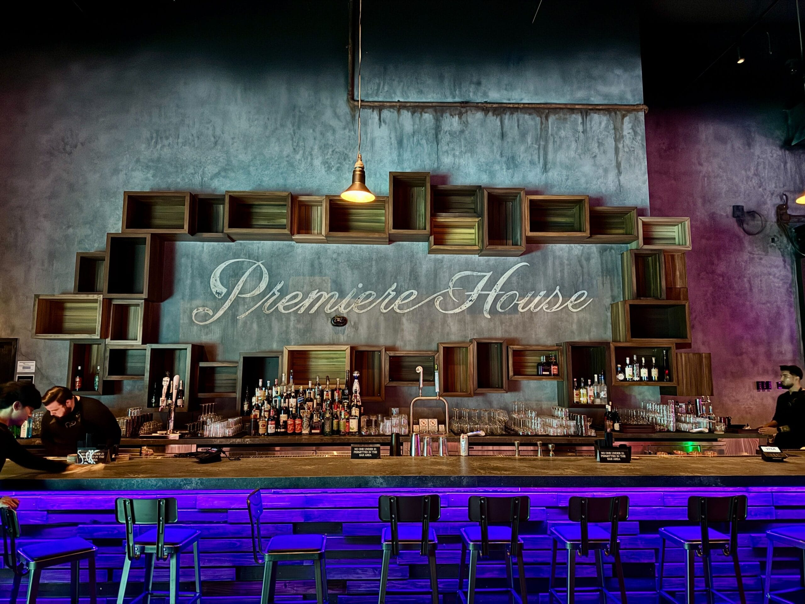 A modern Las Vegas bar with a neon "Première House" sign, shelves of bottles, and empty stools along the counter.
