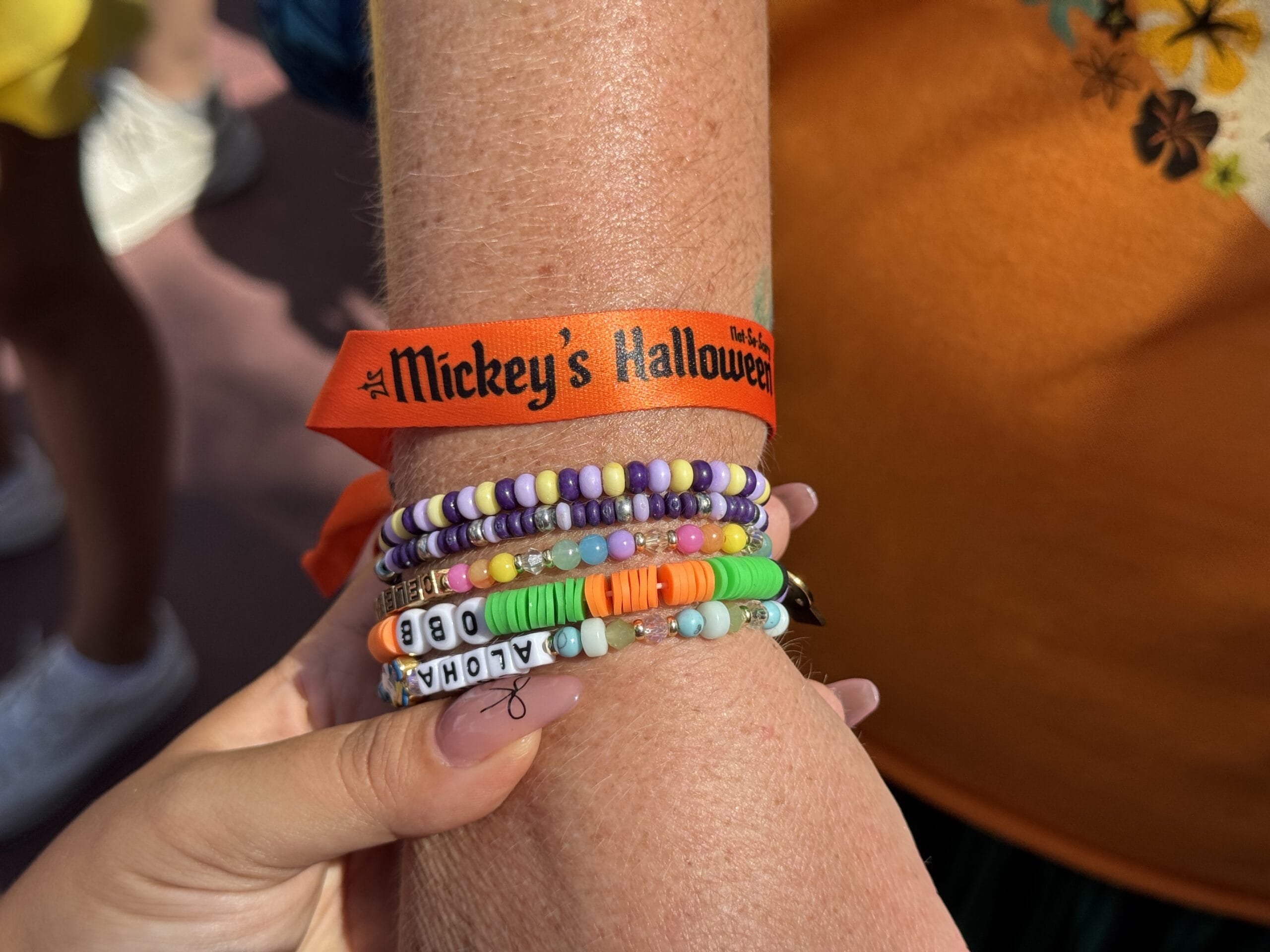 A hand wearing colorful beaded bracelets and an orange Oogie Boogie Bash wristband, held by another hand.