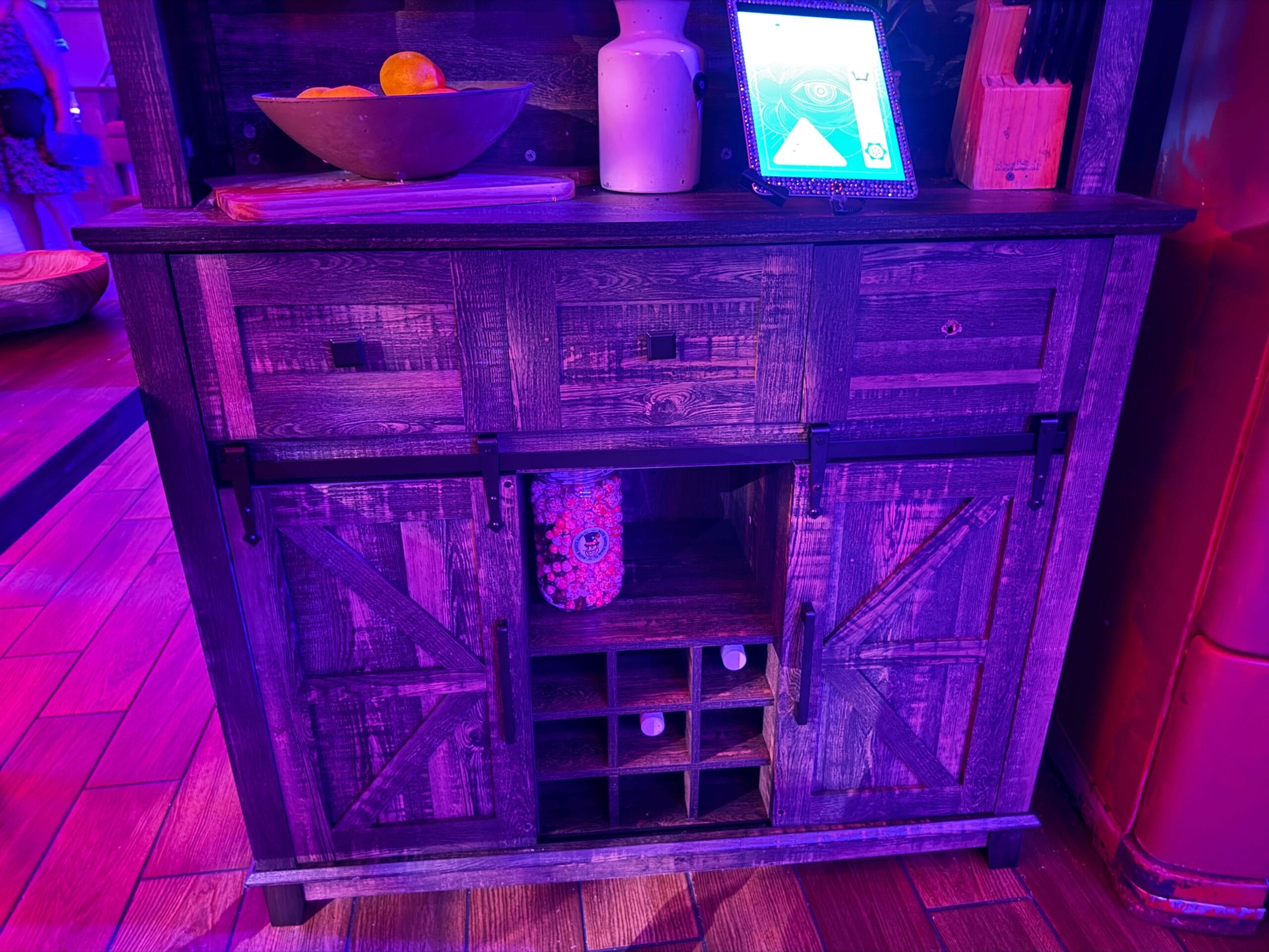 A rustic wooden cabinet with fruit bowl and tablet glows purple-blue—like the Dead Coconut Club on a Sorority Witches full tour.