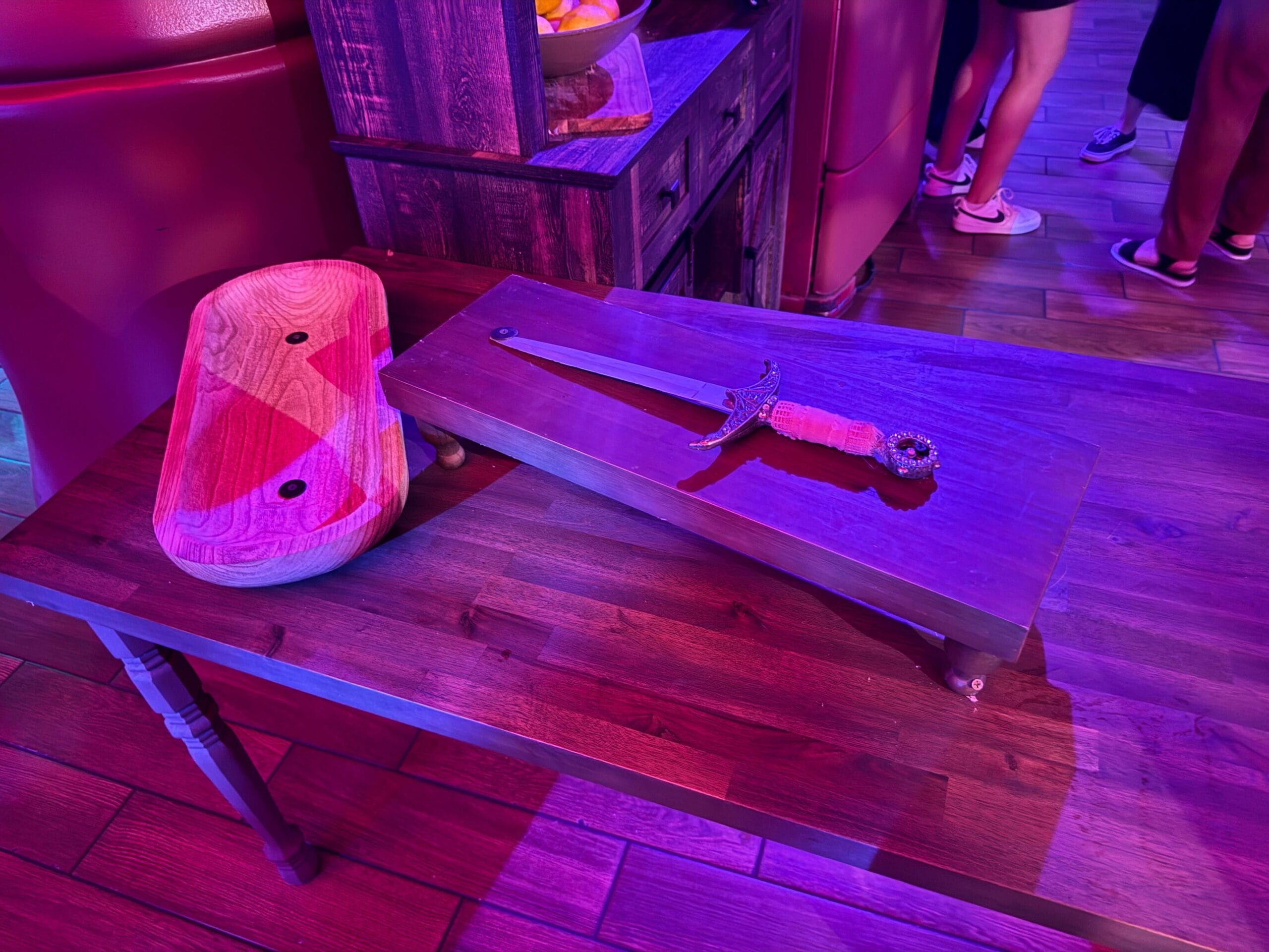 A dagger and wooden dish sit on a table at Dead Coconut Club under purple lighting, with Sorority Witches nearby.