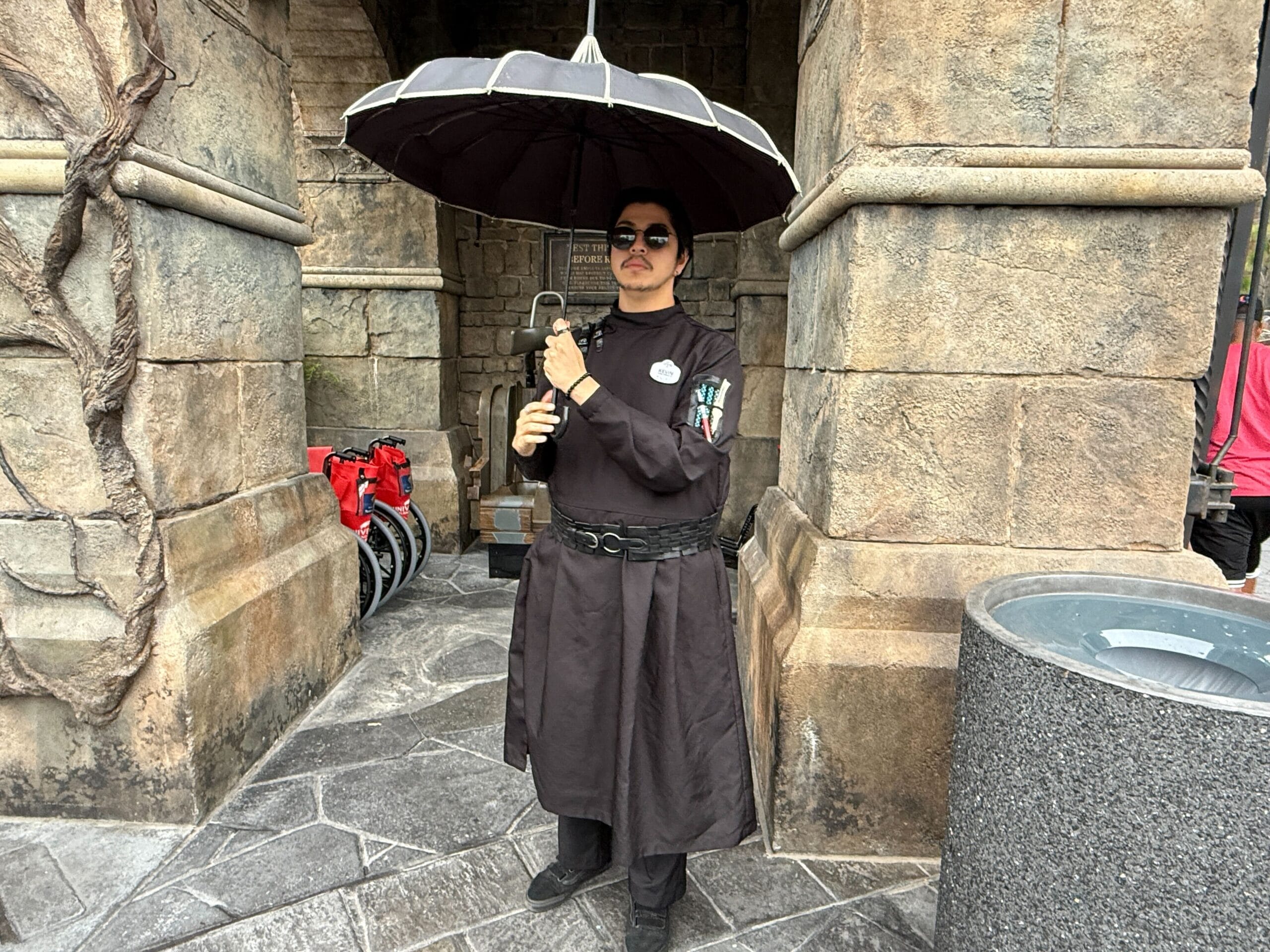 Person in a long dark robe holding a large black umbrella, standing near a stone wall at a themed location.