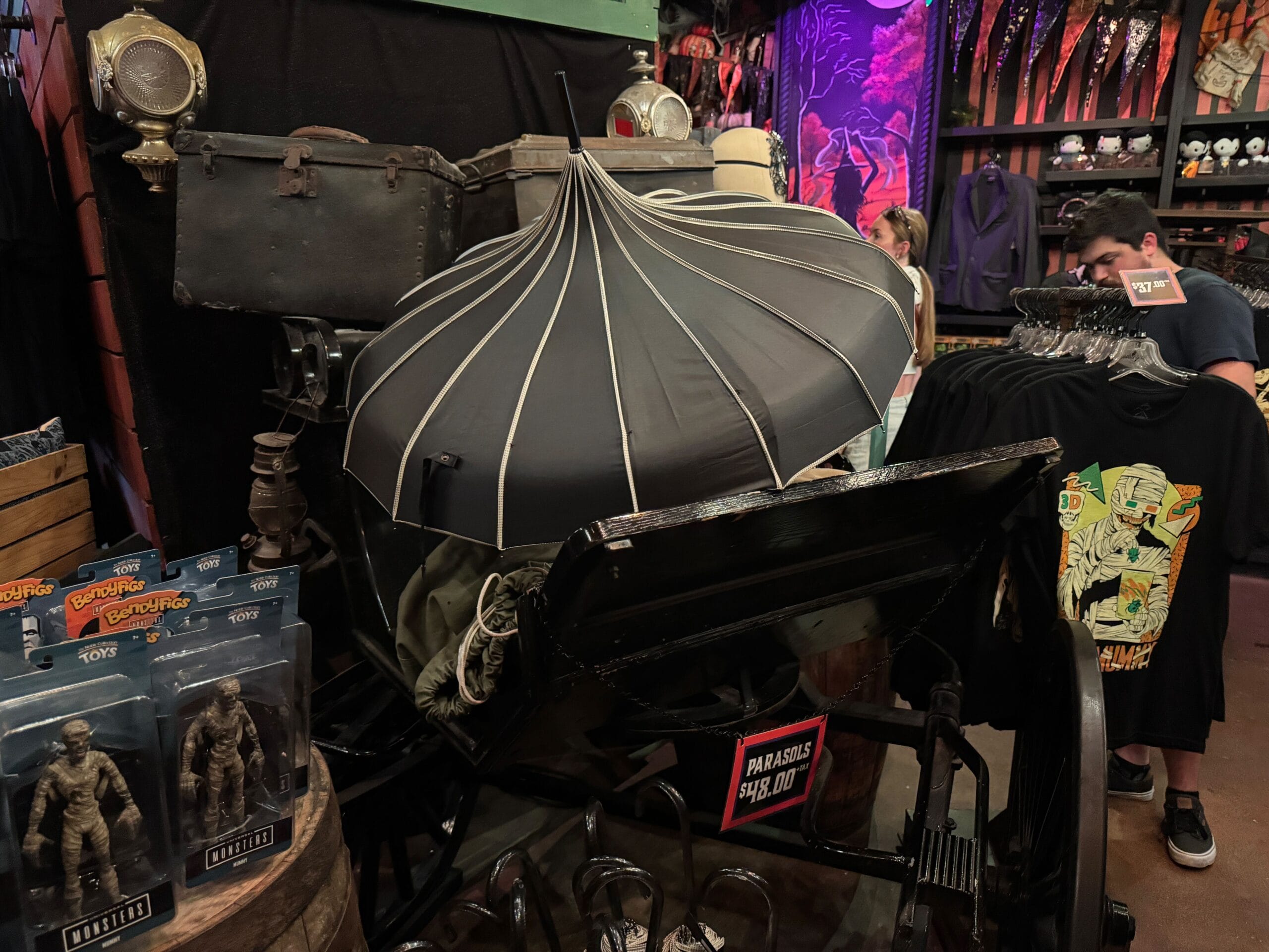 A black parasol shades a vintage stroller in a crowded store selling horror-themed merchandise and action figures.