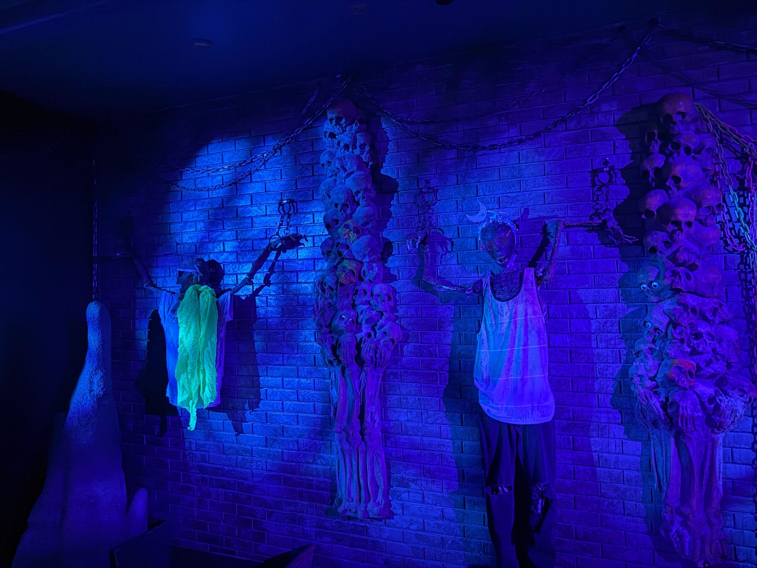 A dimly lit brick wall with chained skeletons and skulls, glowing in blue and purple at the Dead Coconut Club.