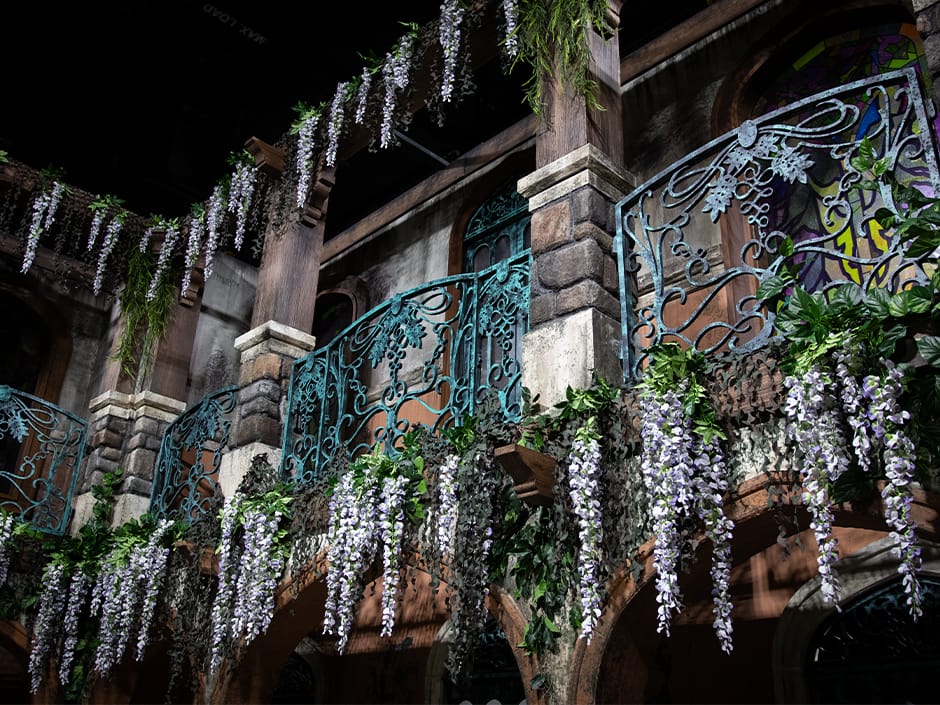 Ornate balcony with teal iron railings and cascading purple wisteria, evoking an El Artista Spanish Haunting.