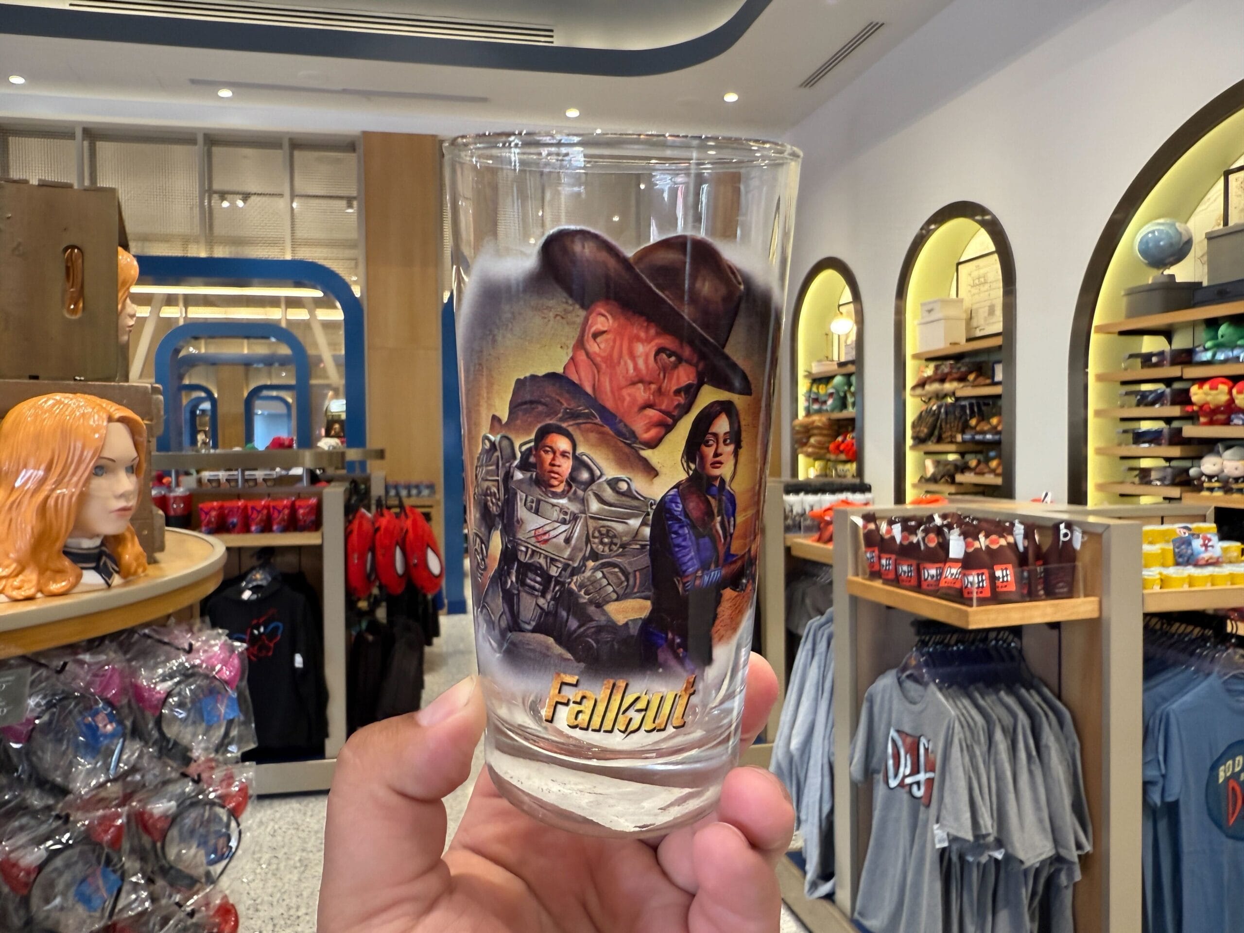 A hand holds a Fallout-themed glass with characters, inside a Universal Orlando Resort store selling T-shirts.