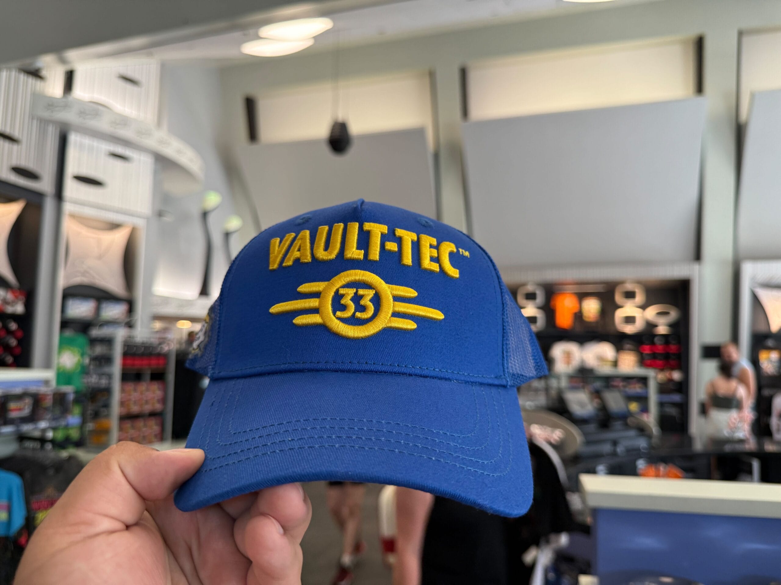 A hand holds a blue "Vault-Tec 33" cap amid Halloween Horror Nights Merchandise at Universal Orlando Resort.