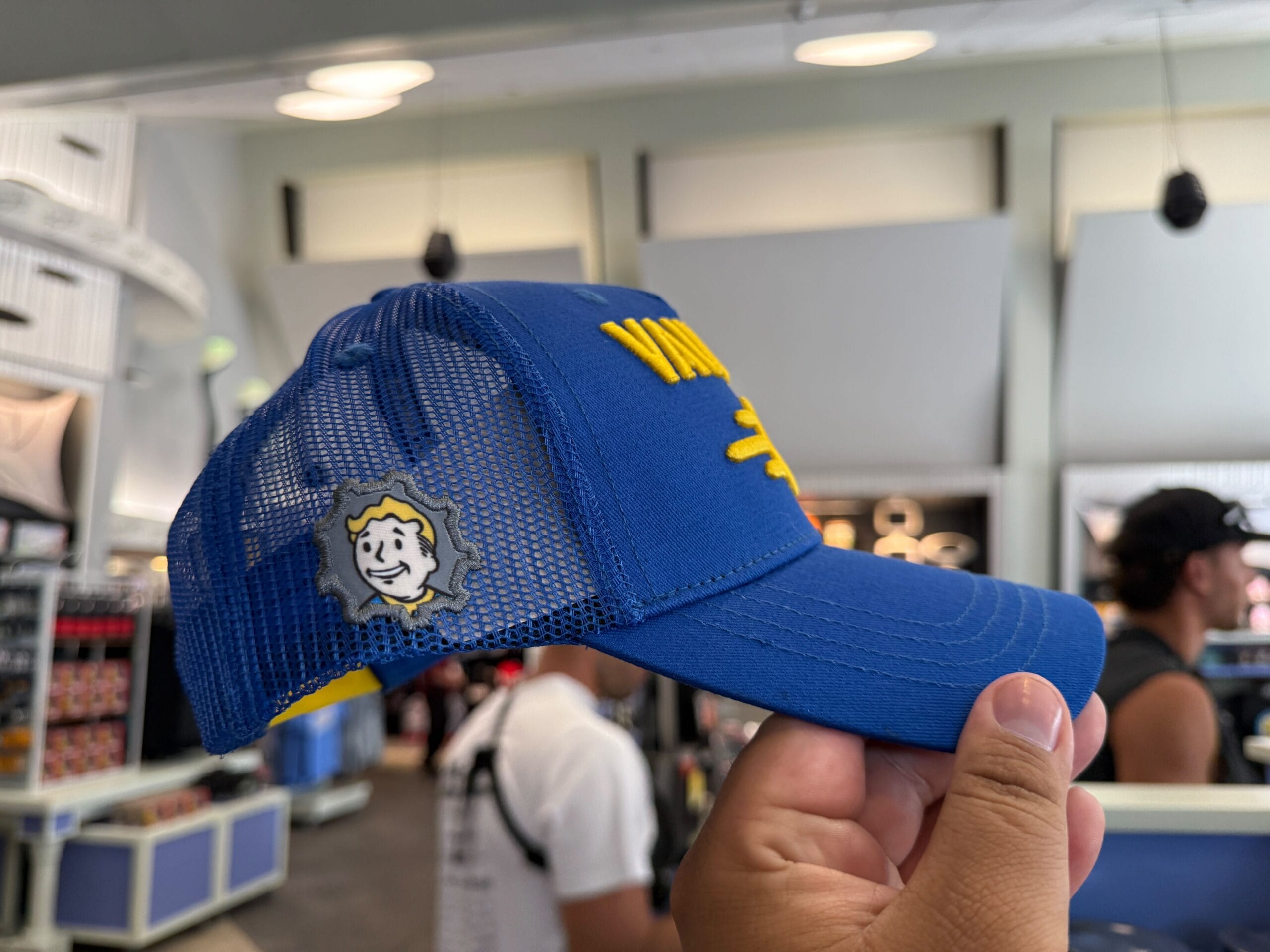 Person holding a blue mesh hat with Vault Boy patch, shopping for Halloween Horror Nights Merchandise at Universal Orlando Resort.