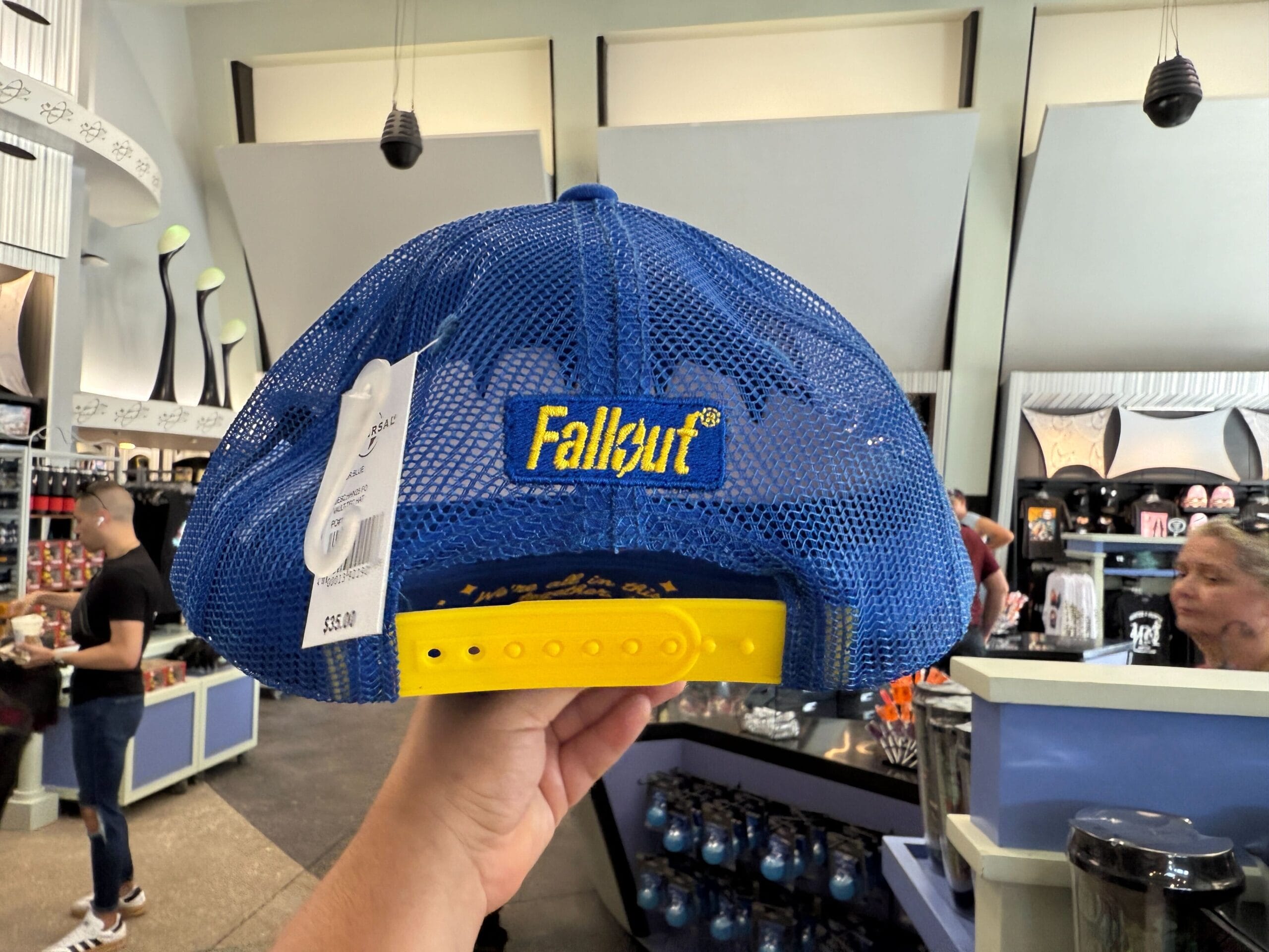 A hand holds a blue mesh "Fallout" cap with a yellow strap, spotted among HHN Merch 2025 at Universal Orlando Resort.