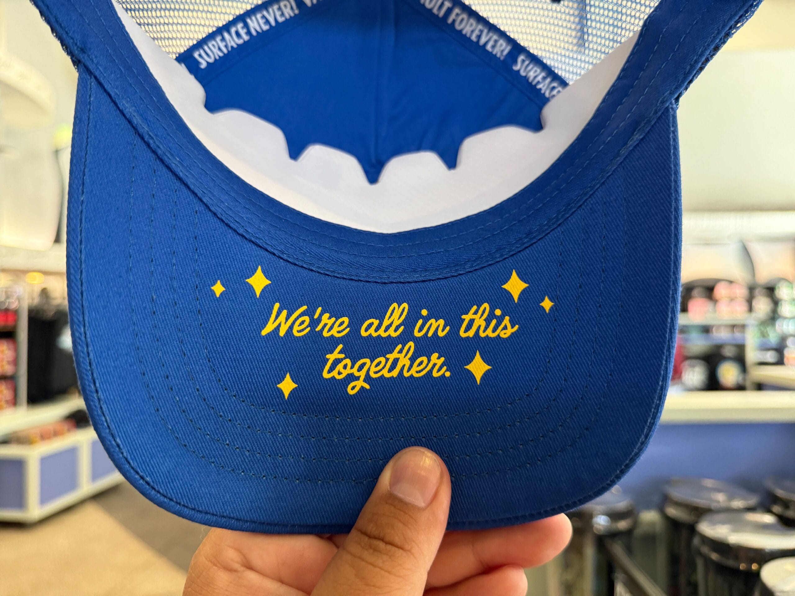 A blue hat brim with “We’re all in this together” in yellow, held by a hand—perfect for Universal Orlando Resort fans.