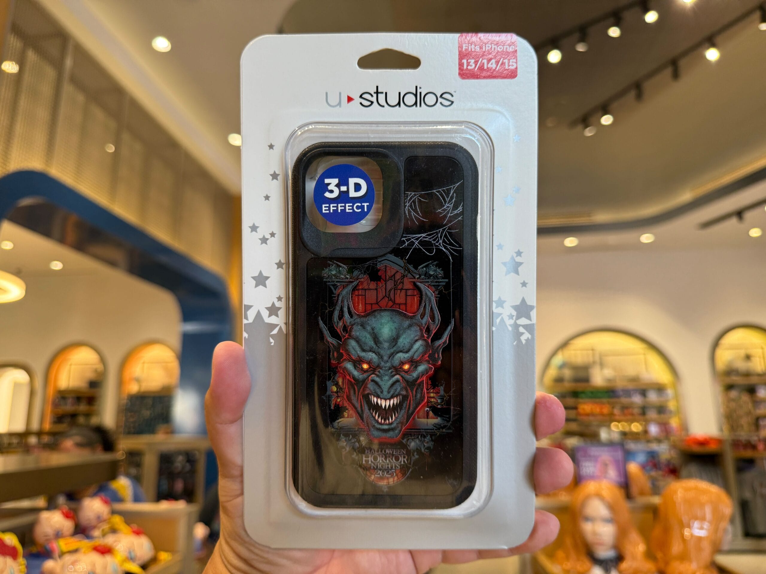 A hand holds packaged HHN Merch 2025 with a red demon face and "3-D effect" label inside Universal Orlando Resort store.