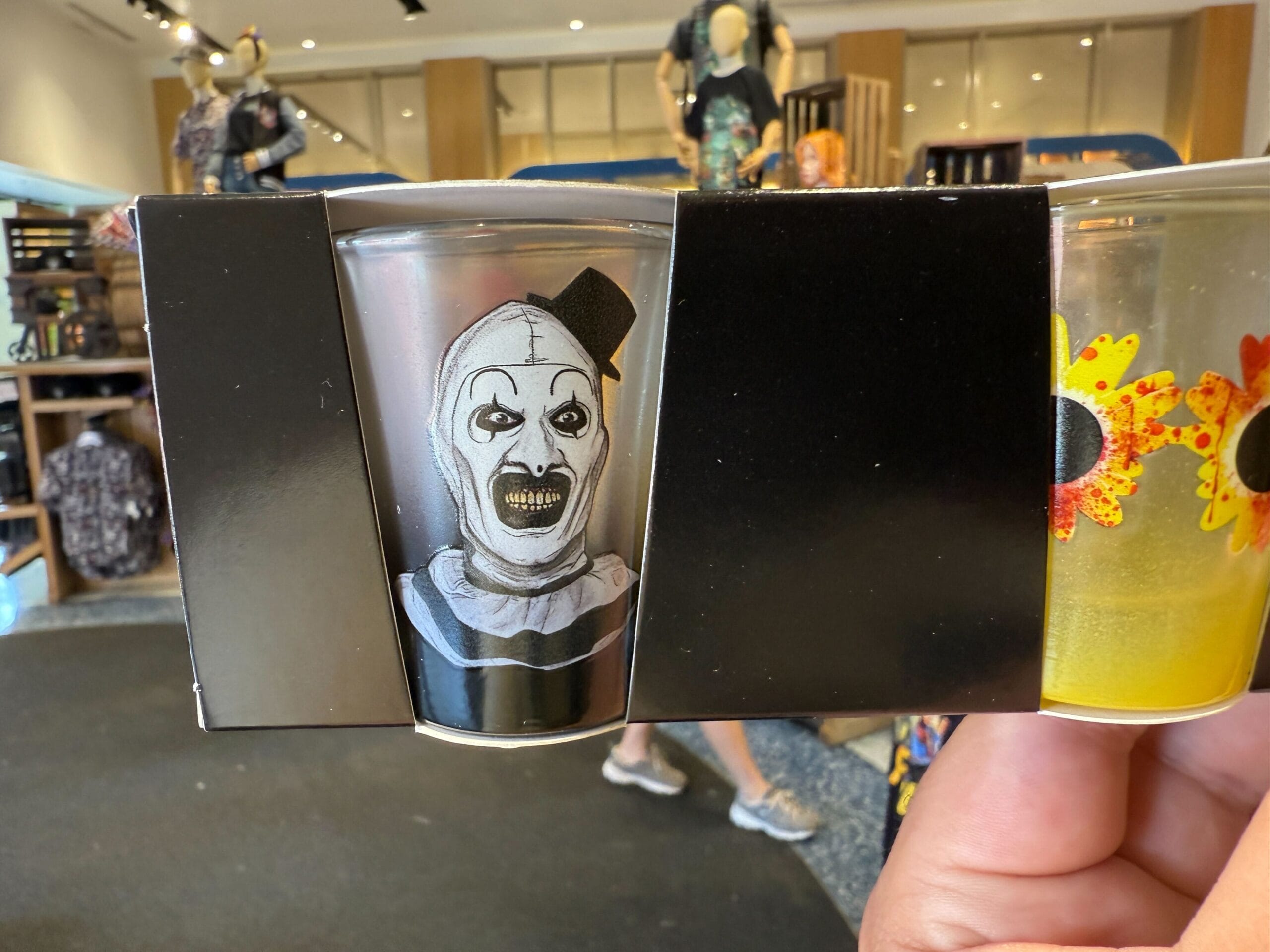A hand holds a shot glass featuring a scary clown face—perfect for Halloween Horror Nights Merchandise collectors.