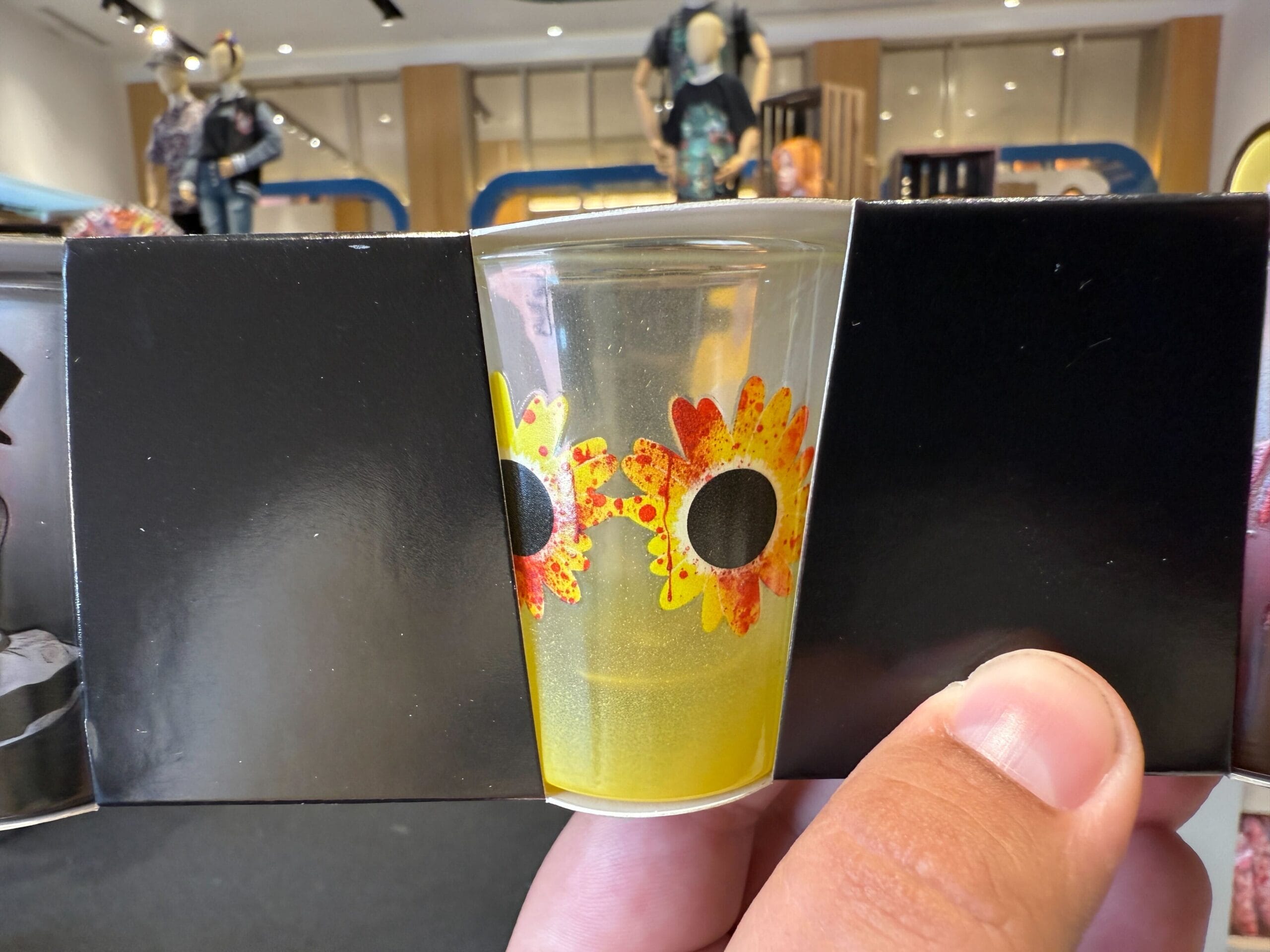 A clear cup with sunflower designs, part of HHN Merch 2025, is held between two black panels like eyes and sunglasses.