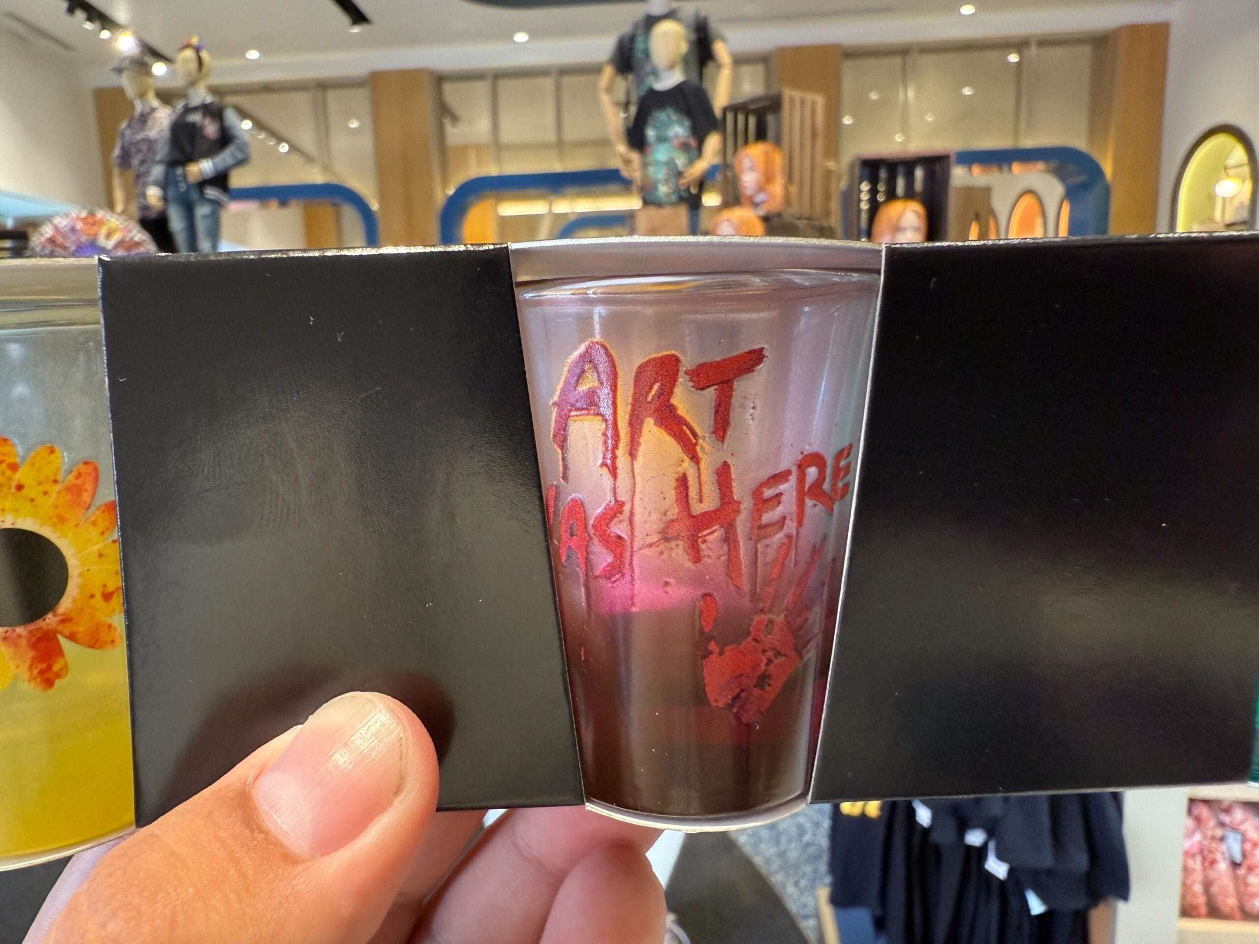 A hand holds a glass reading "ART WAS HERE" in red, displayed with Halloween Horror Nights Merchandise in the background.