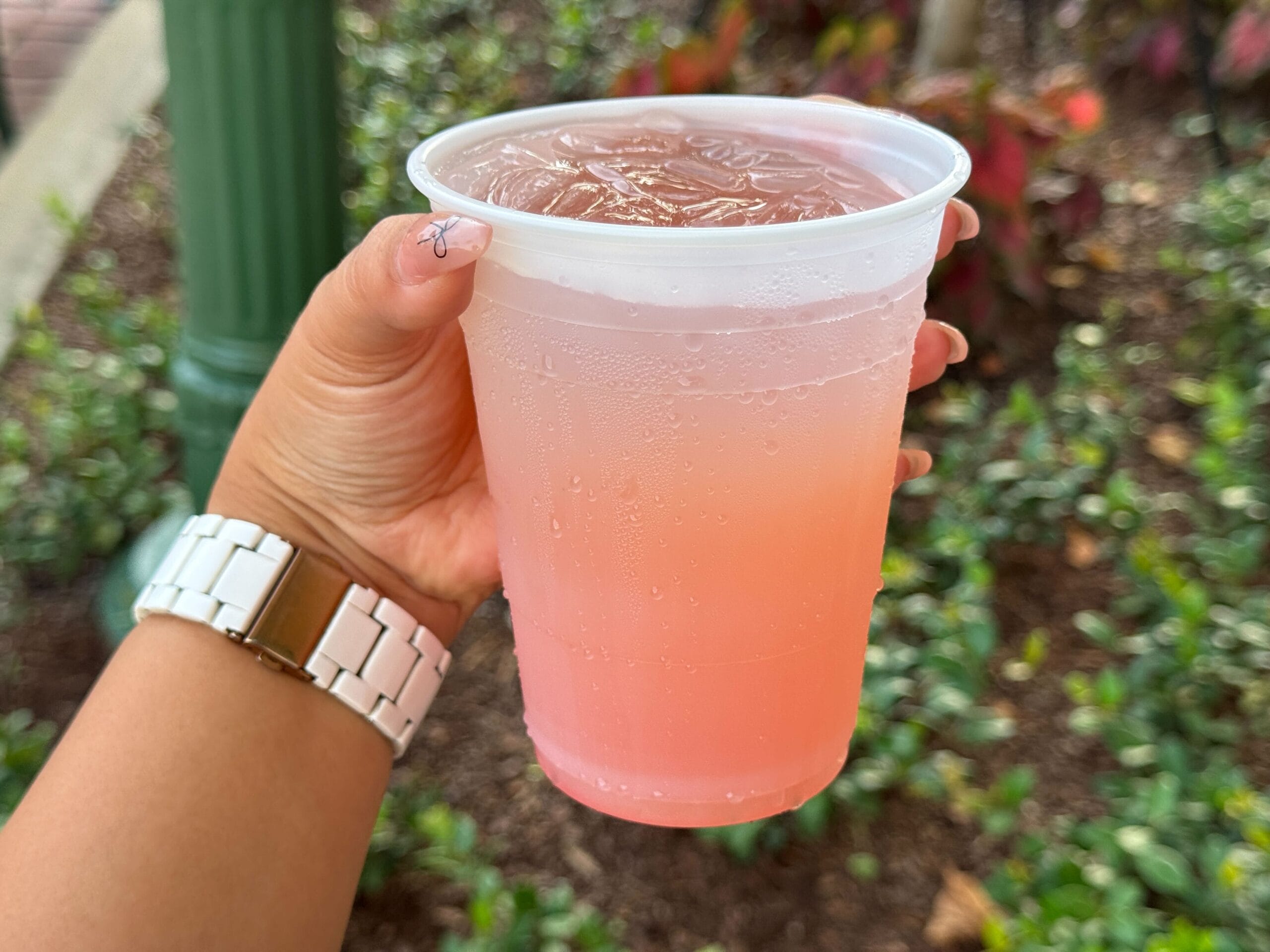 A hand holds a plastic cup filled with a pink iced drink outdoors, with plants and a green post in the background.
