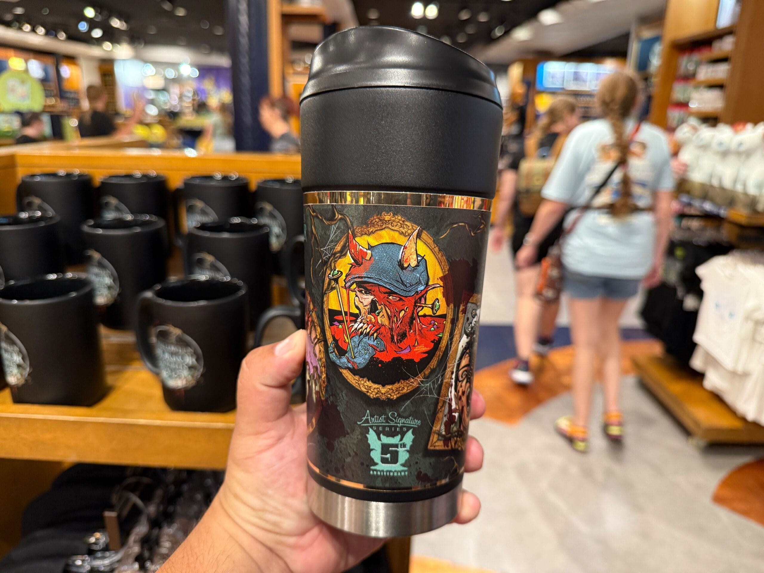 A hand holds Halloween Horror Nights Merchandise at Universal Orlando Resort, with black mugs and shoppers in the background.