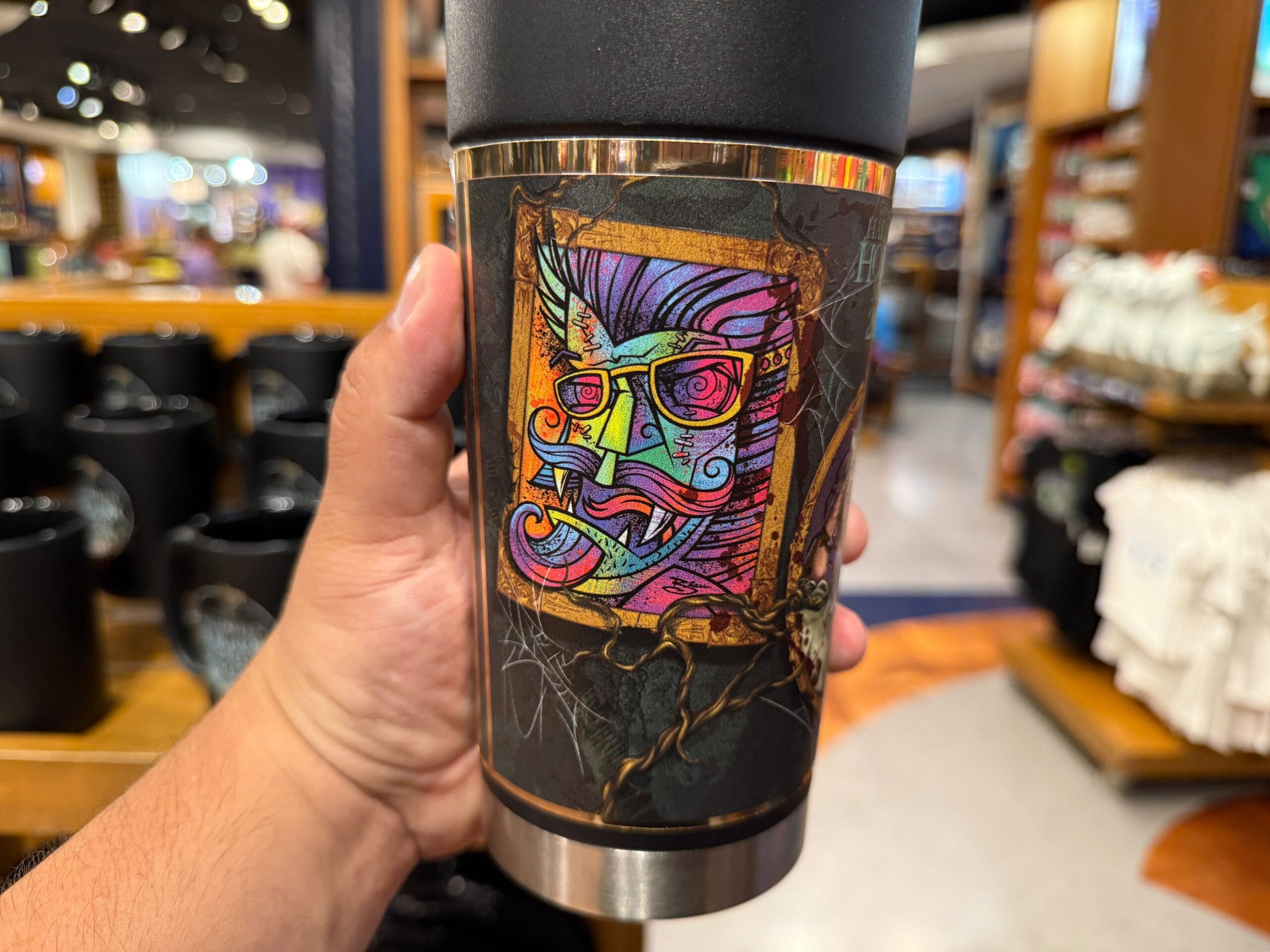 A hand holds a tumbler with a colorful, abstract face amid shelves of HHN Merch 2025 at Universal Orlando Resort.