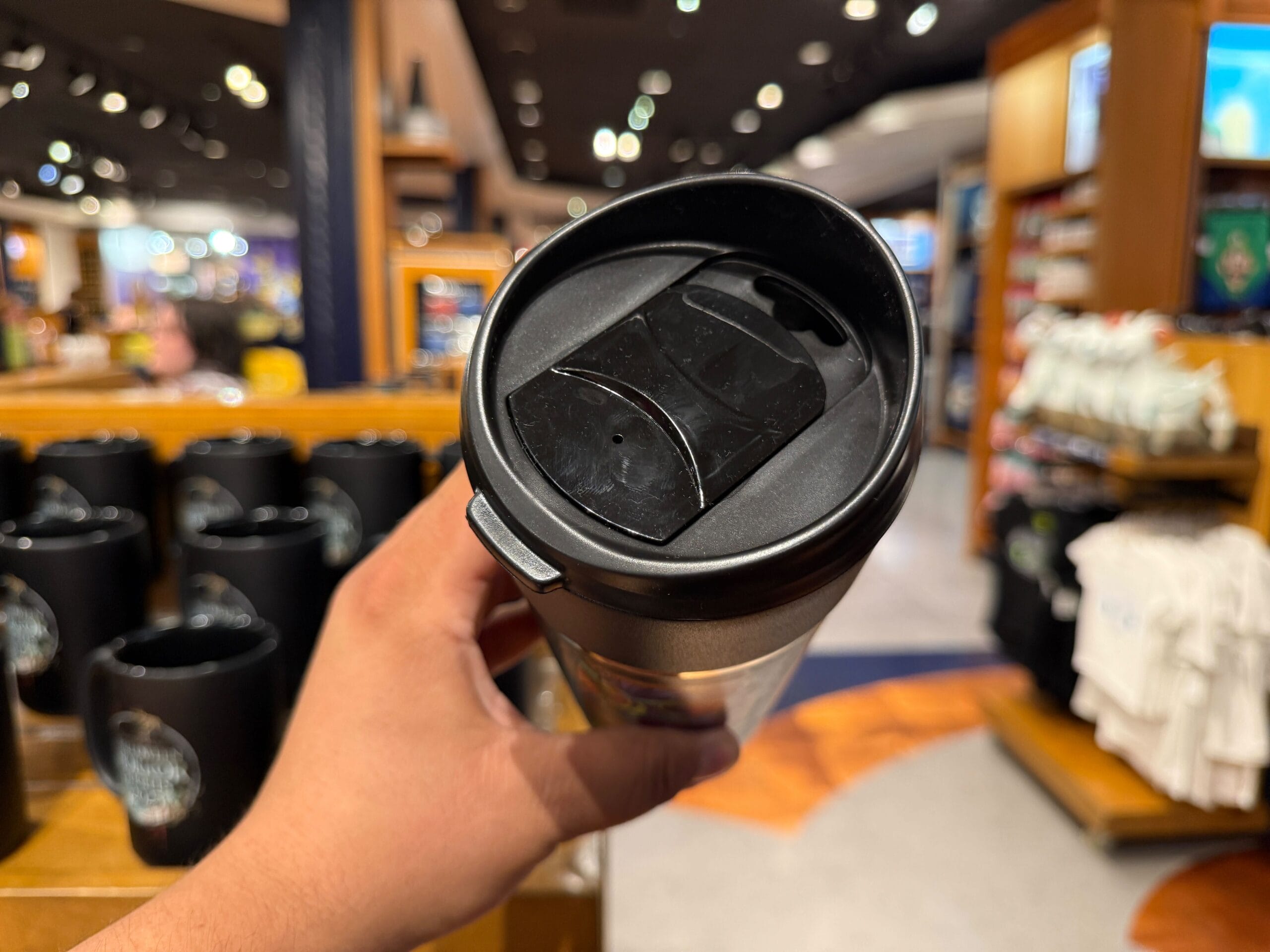 A hand holds a black travel mug in a store, surrounded by blurred Halloween Horror Nights Merchandise in the background.