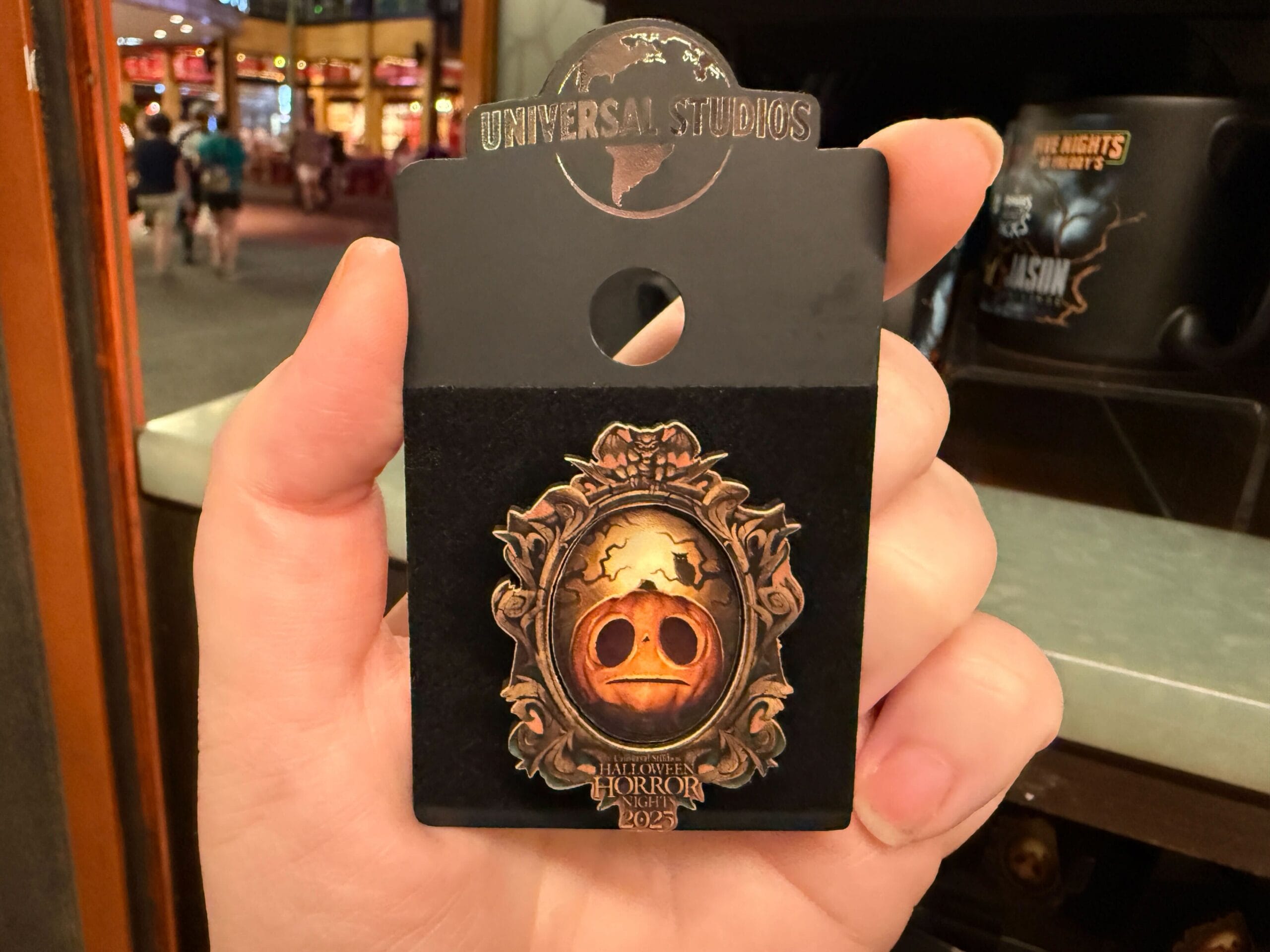 Hand holding a Universal Studios Halloween Horror Nights Merchandise pin with a carved pumpkin face in an ornate frame.