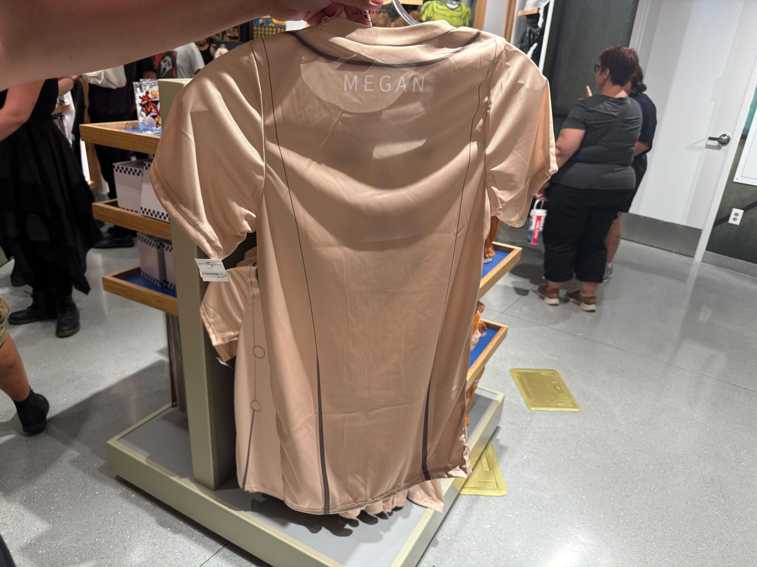 A person displays a beige "MEGAN" shirt, part of Halloween Horror Nights Merchandise at Universal Orlando Resort.