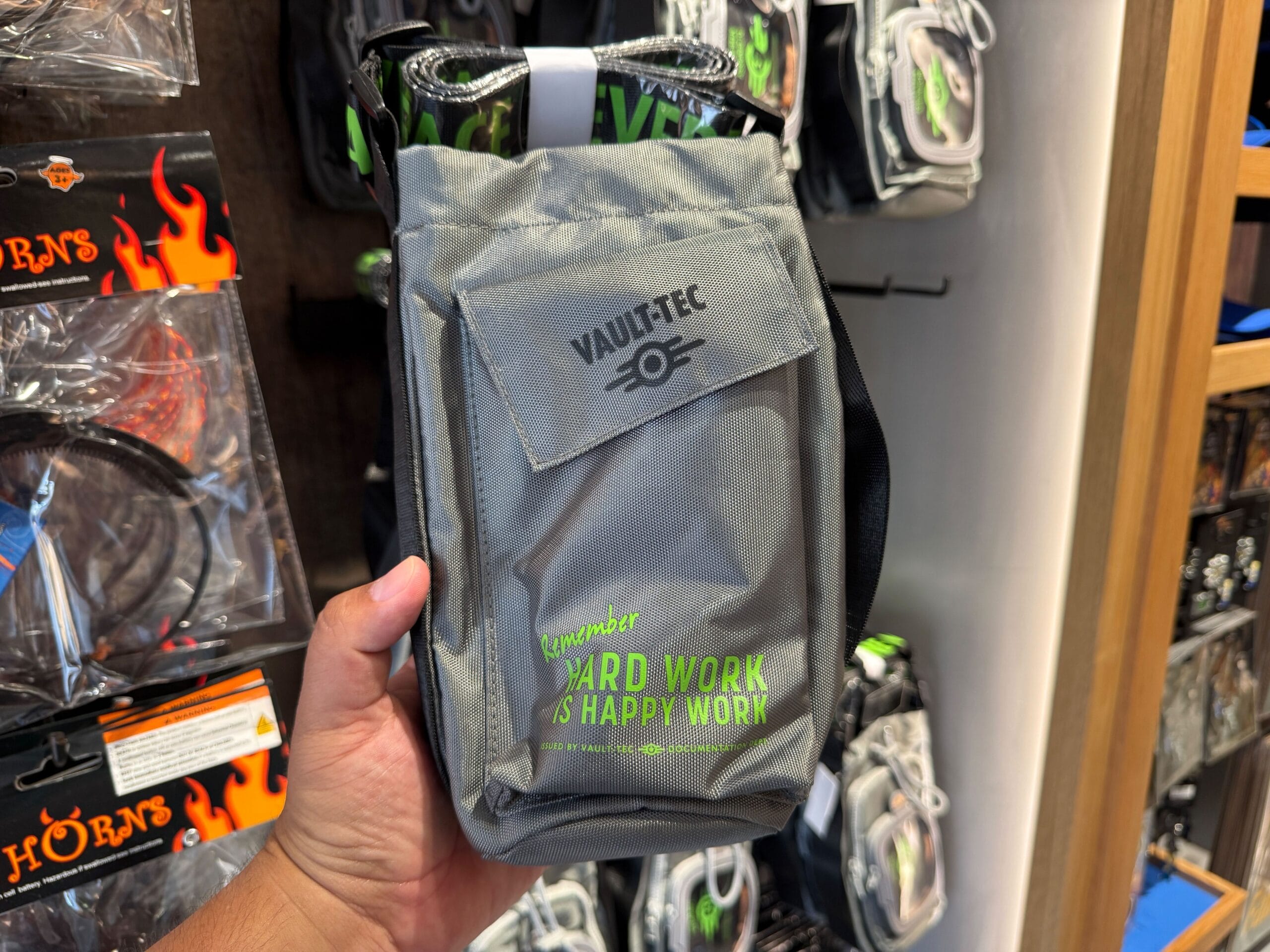 Hand holding a gray Vault-Tec pouch from Universal Orlando Resort, store display reads: "Remember: Hard Work is Happy Work.