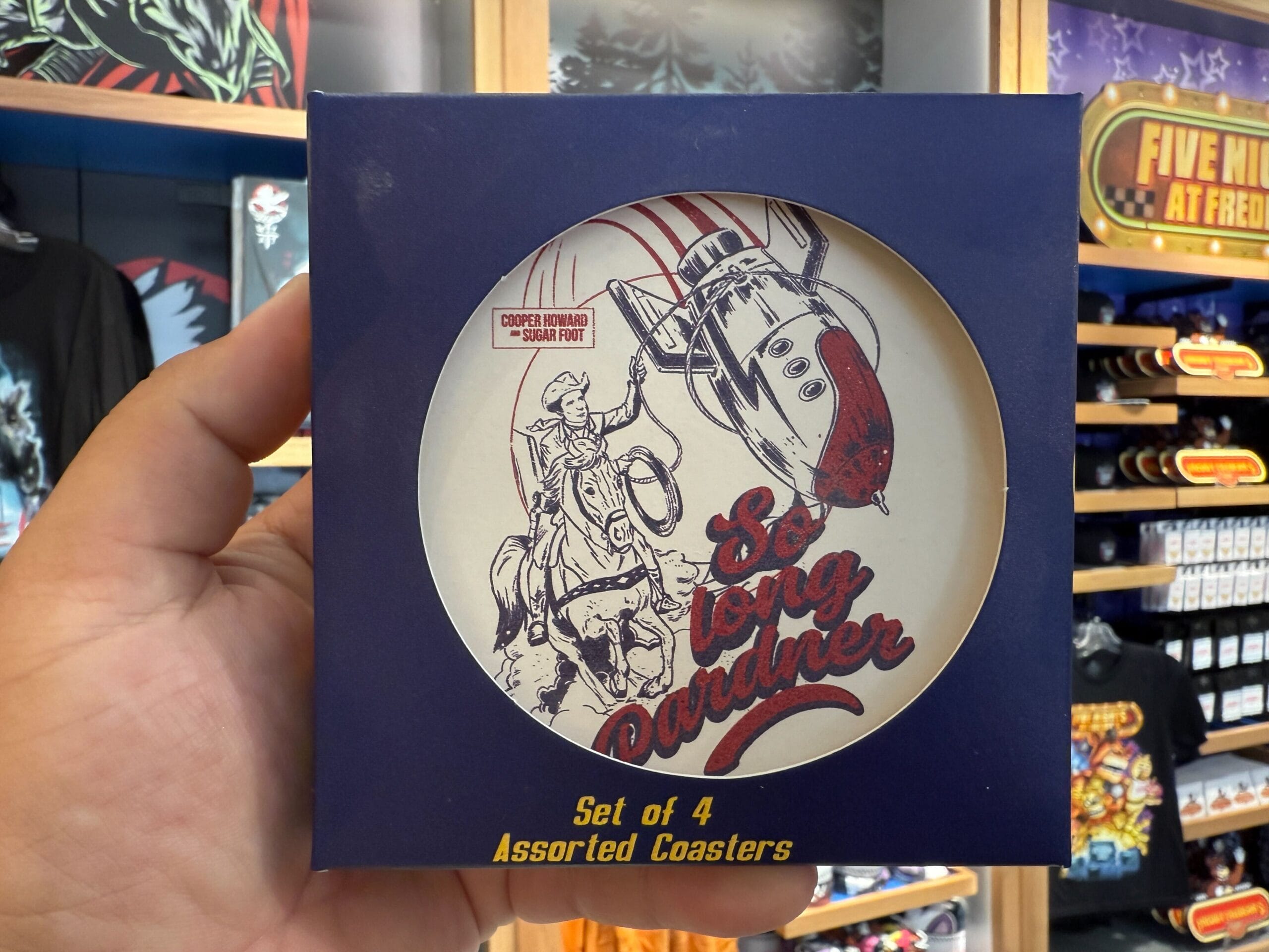 A hand holds a box of coasters with a cowboy, rocket, and "So long pardner." Exclusive HHN Merch at Universal Orlando Resort.