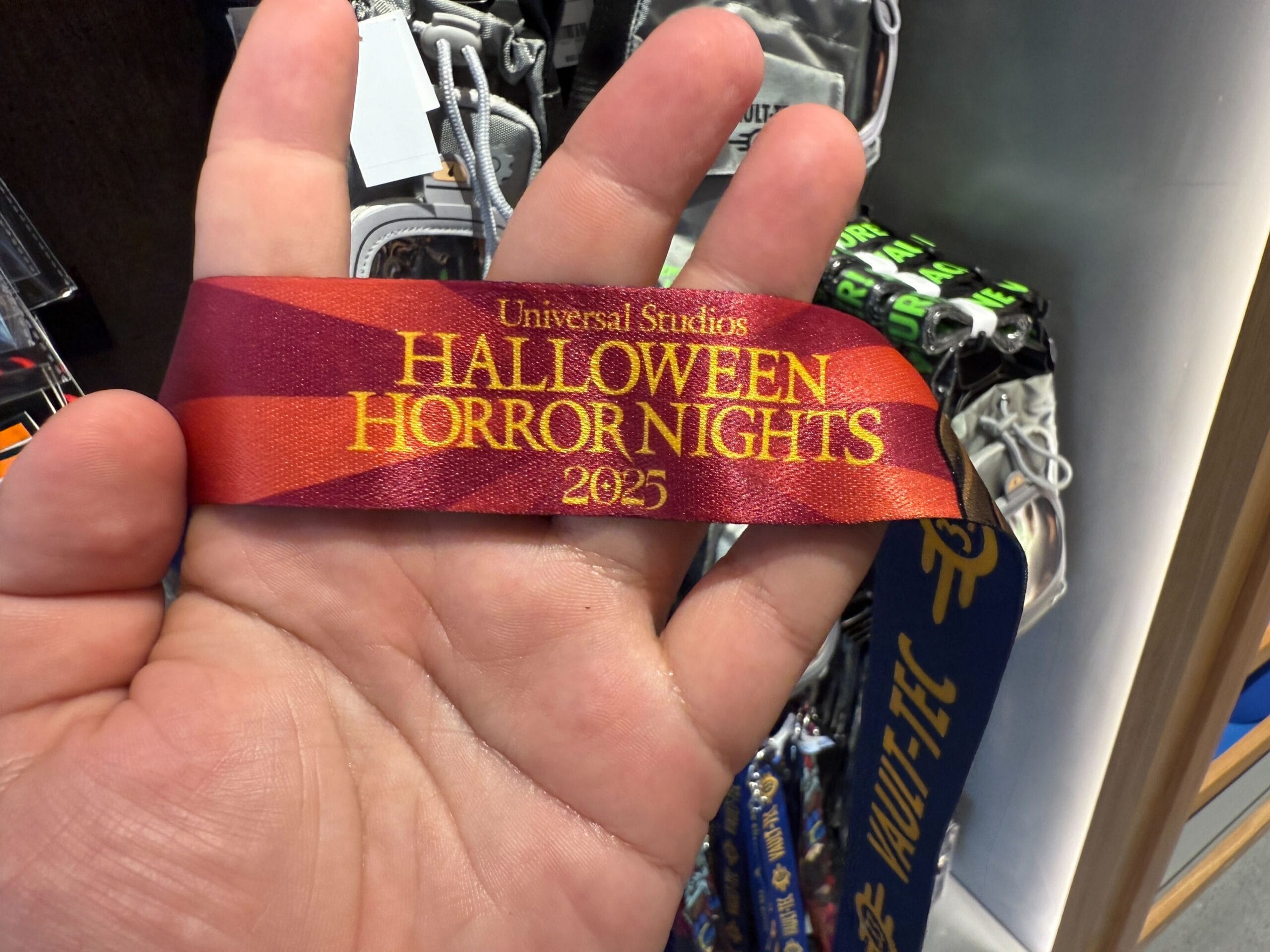 A hand holds a ribbon that reads "Universal Studios Halloween Horror Nights 2025"—a must-have HHN Merch item.