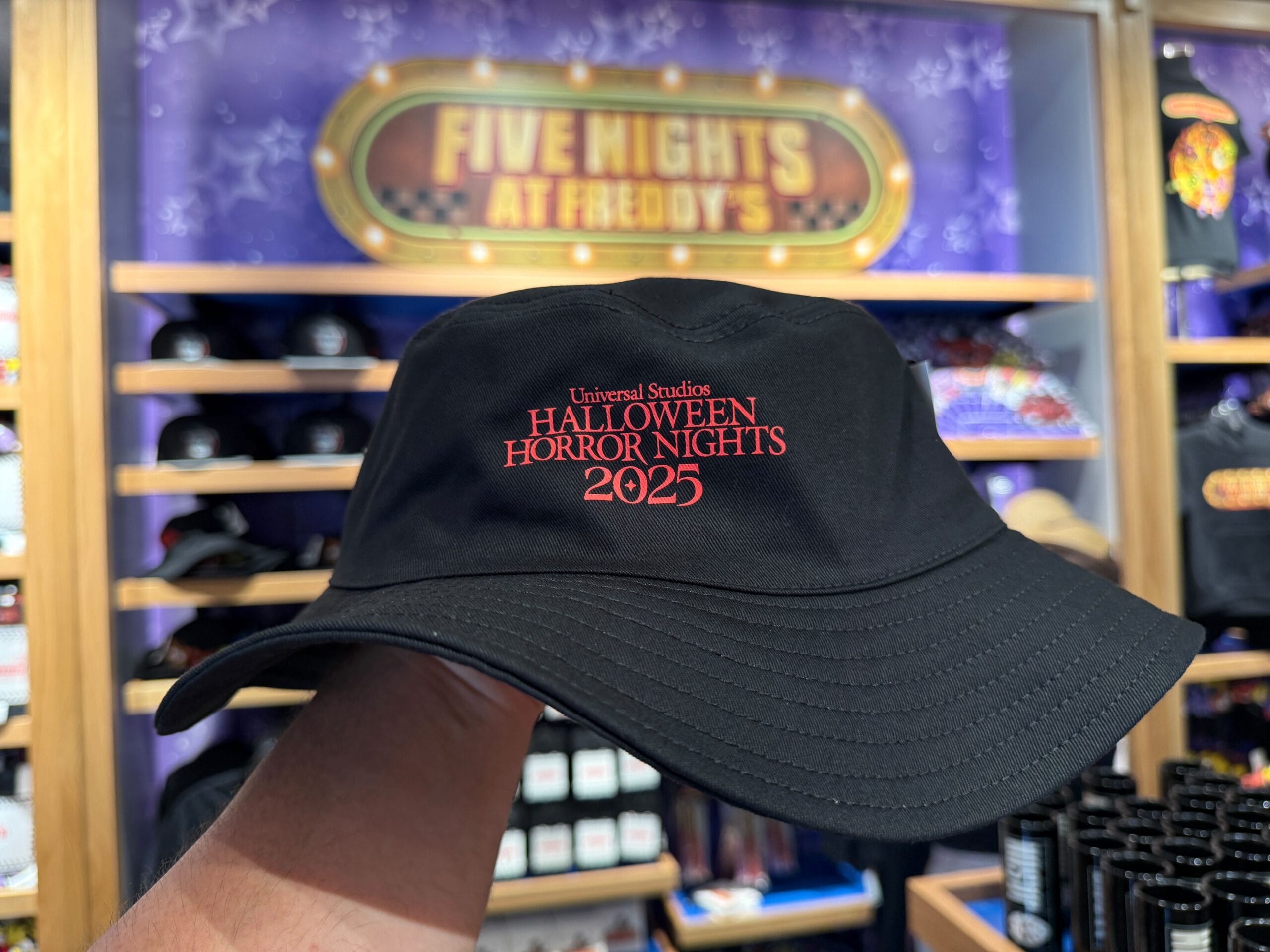 A hand holds an HHN Merch black bucket hat with "Halloween Horror Nights 2025" at a Universal Studios store.