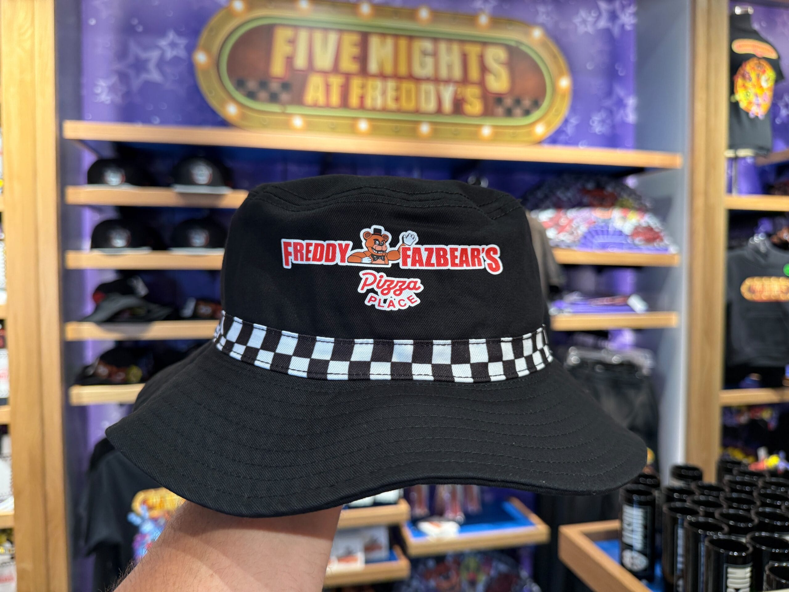 A hand holds a black bucket hat with a Freddy Fazbear's Pizza Place logo, spotted at Universal Orlando Resort HHN Merch store.