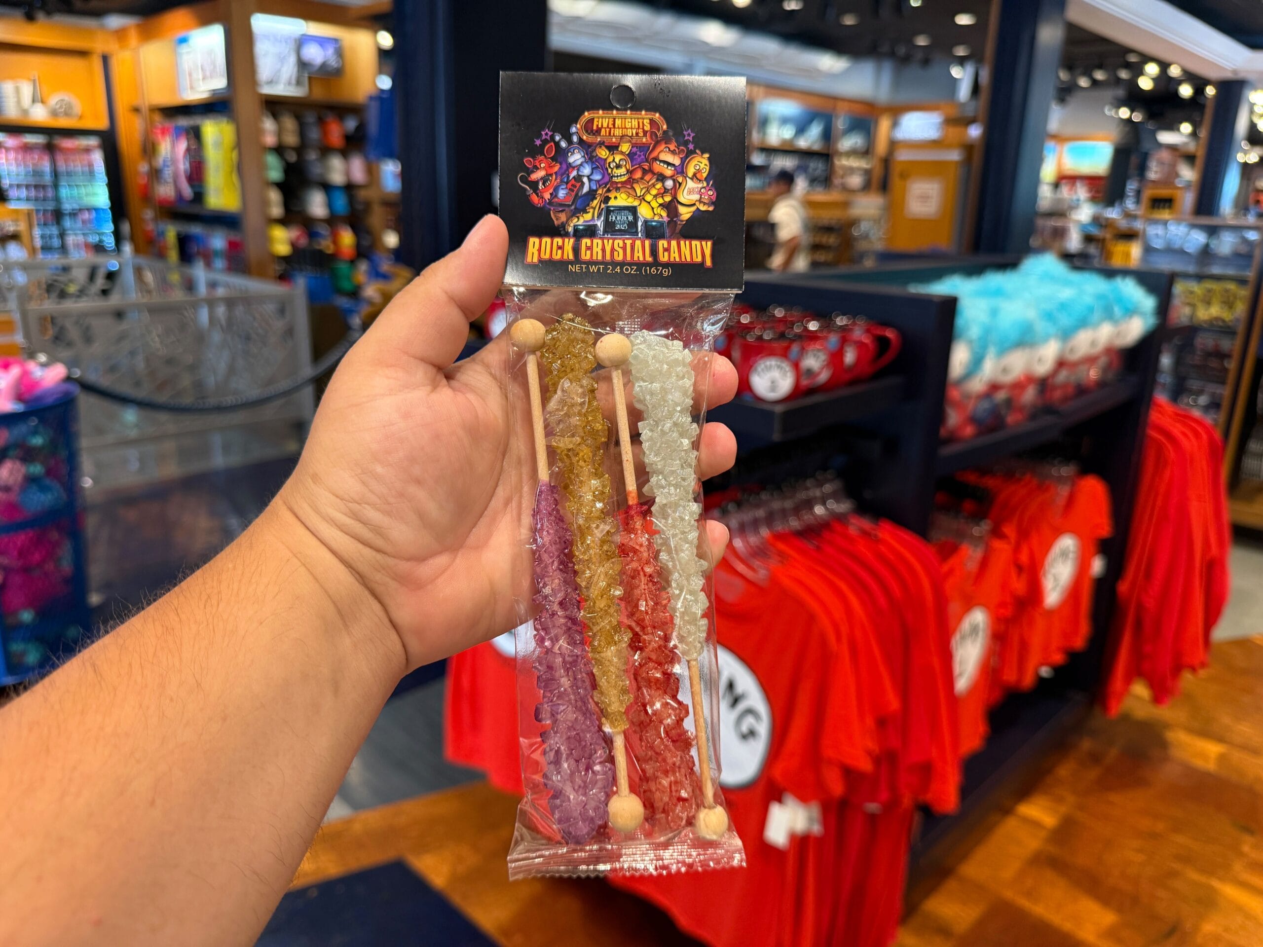 A hand holds three sticks of rock crystal candy in a Universal Orlando Resort store with colorful shirts and HHN Merch behind.