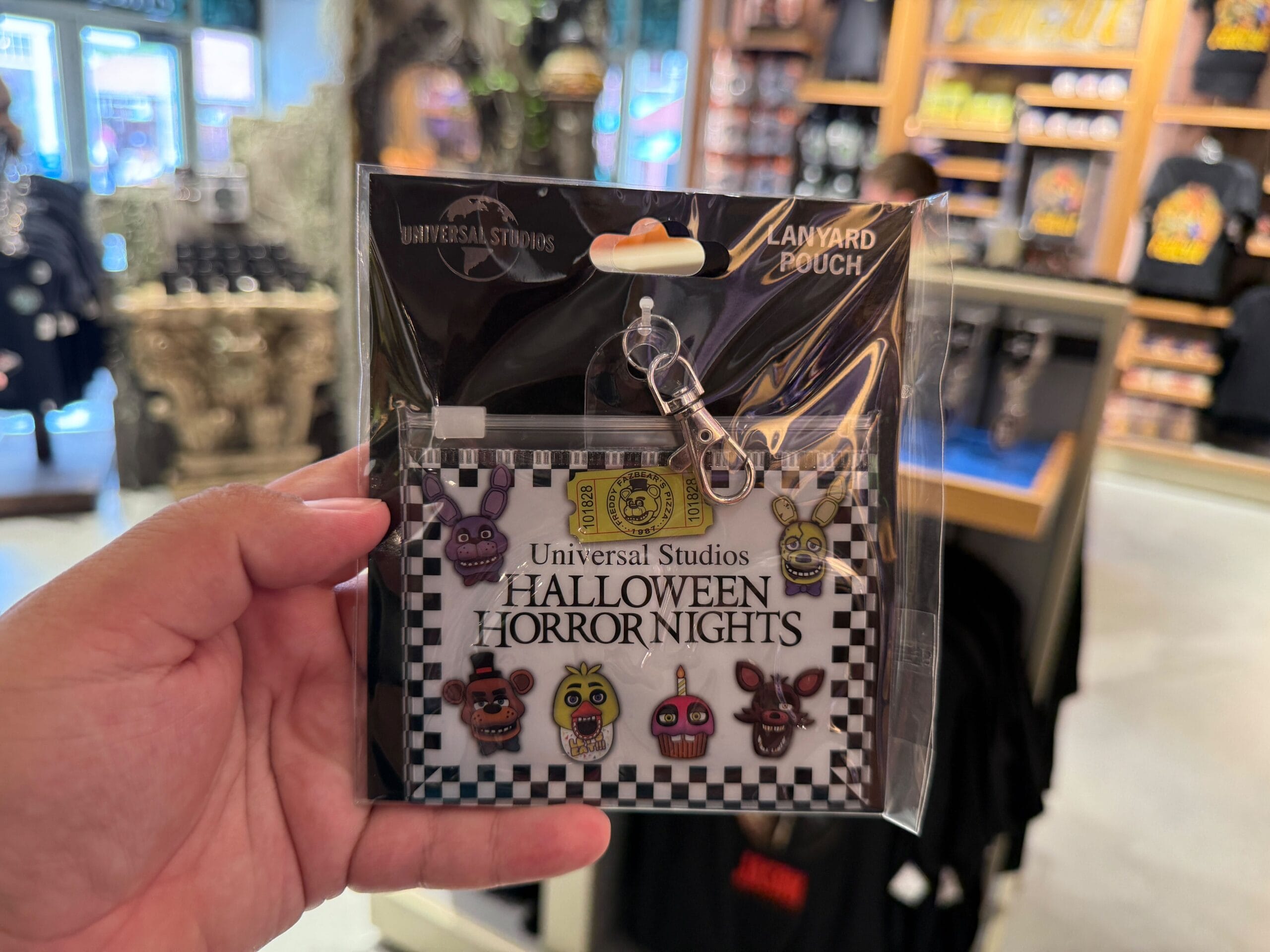 A hand holds HHN merch—a Universal Orlando Halloween Horror Nights lanyard pouch with cartoon monster faces in a store.