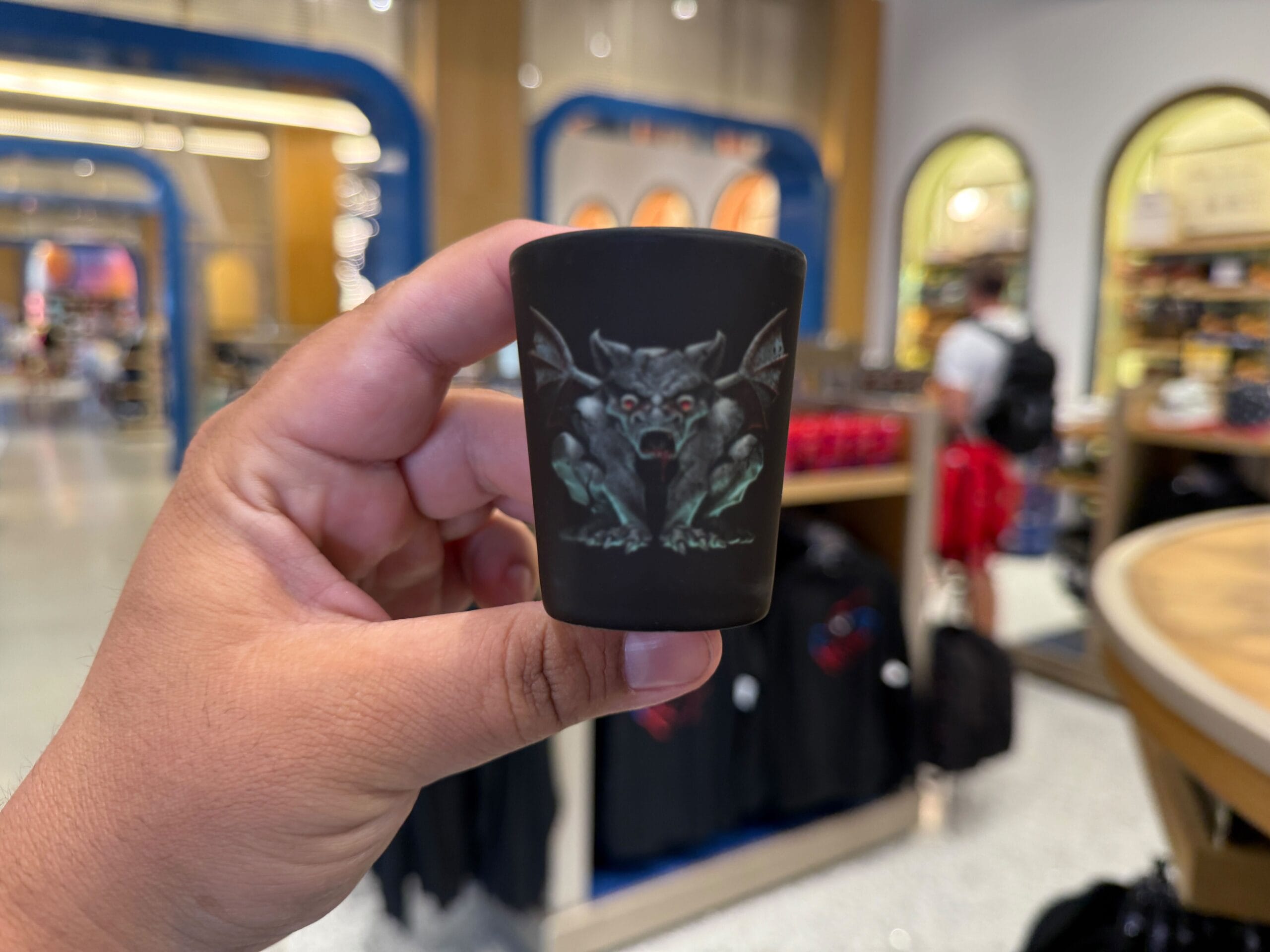 A hand holds a black shot glass with a snarling gargoyle, part of Universal Orlando Resort's HHN Merch in a retail store.