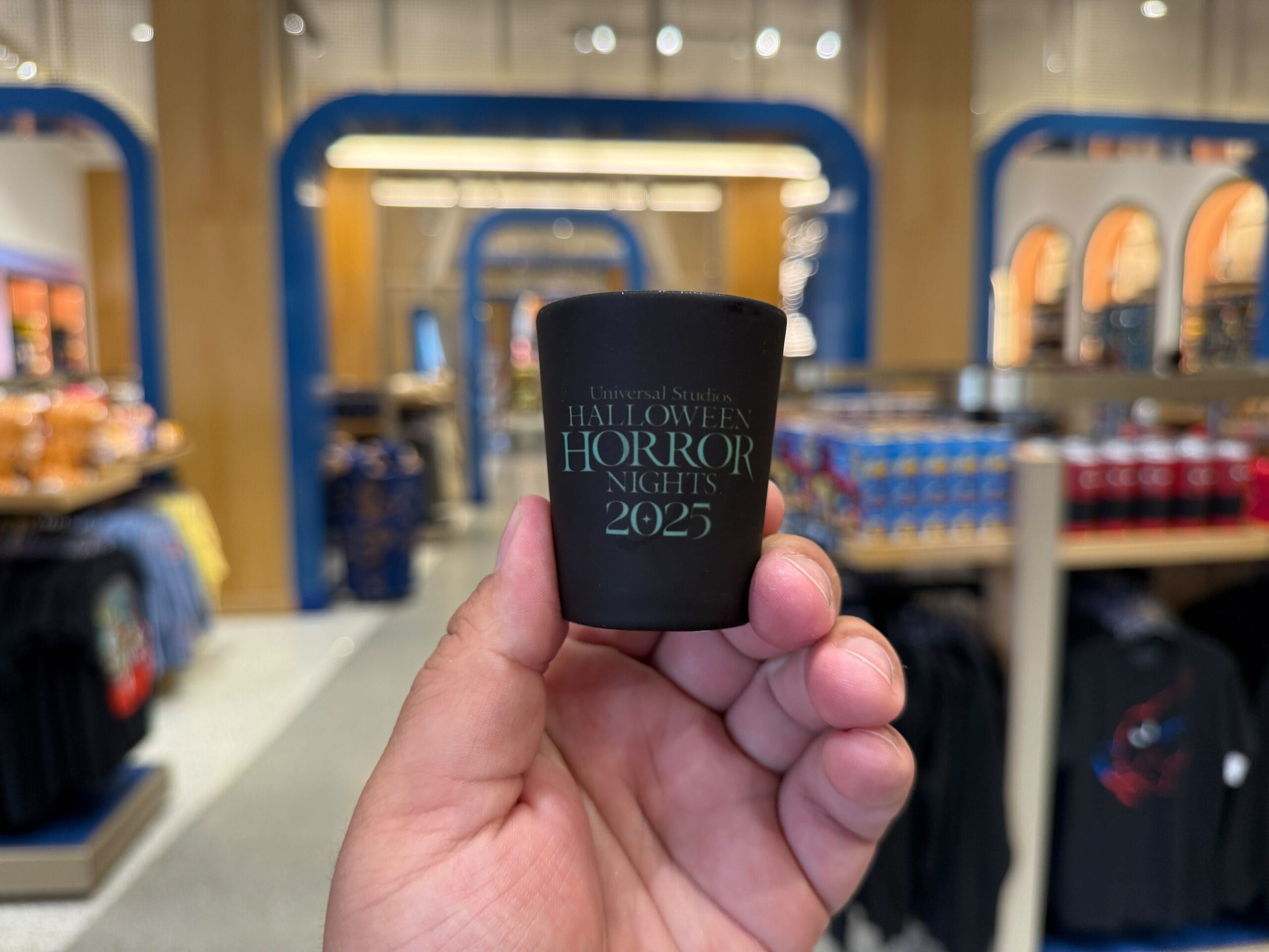 A hand holds a black shot glass labeled “Halloween Horror Nights 2025 Merchandise” at Universal Orlando Resort.