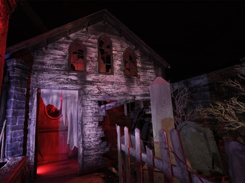 A spooky, abandoned house with broken windows, eerie red lighting, and a graveyard in front.