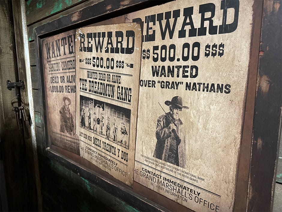 Old-fashioned wanted posters offering rewards and featuring cowboy images hang on a rustic wooden wall.