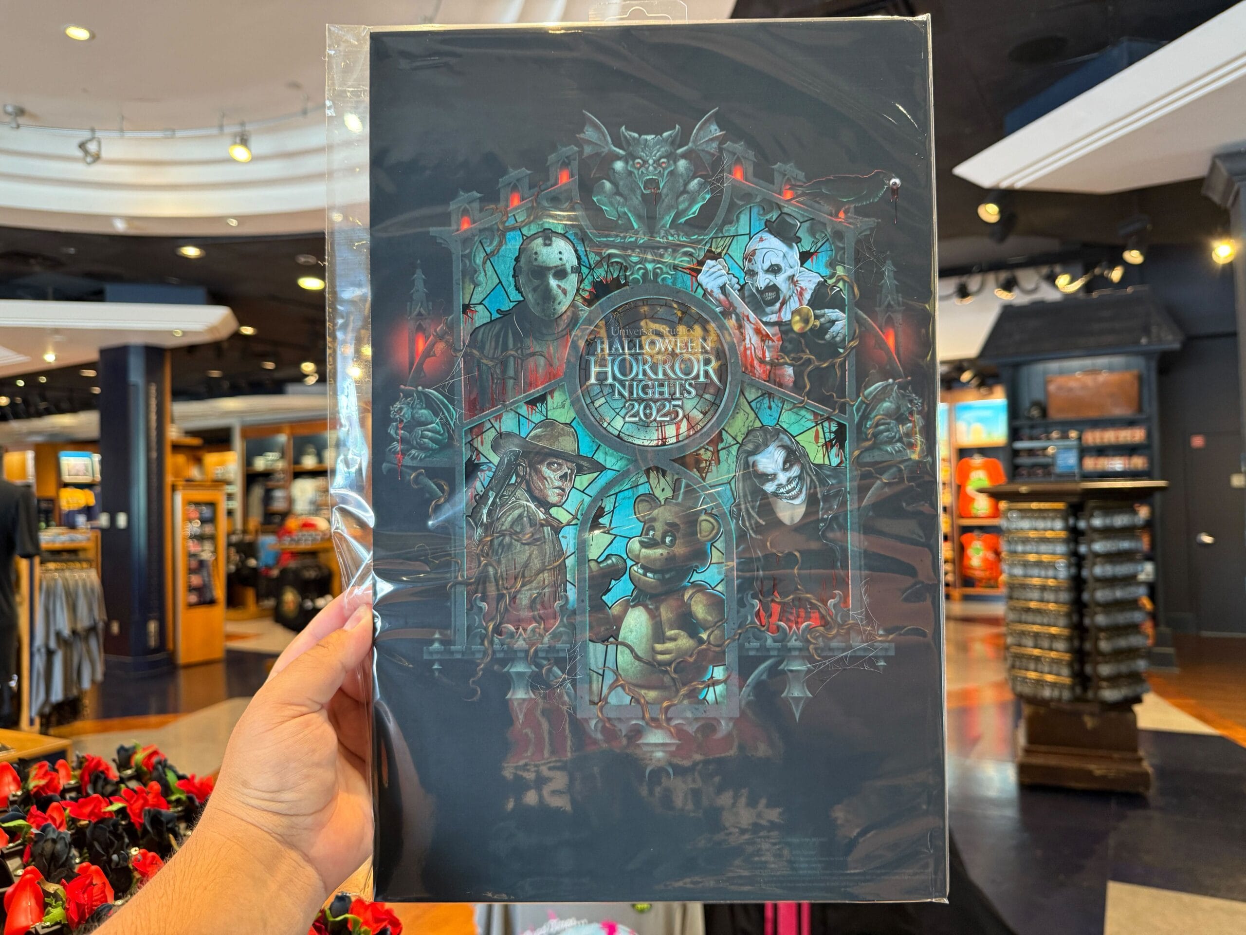 Hand holding a Halloween Horror Nights 2025 poster with horror characters, inside a Universal Orlando Resort store.