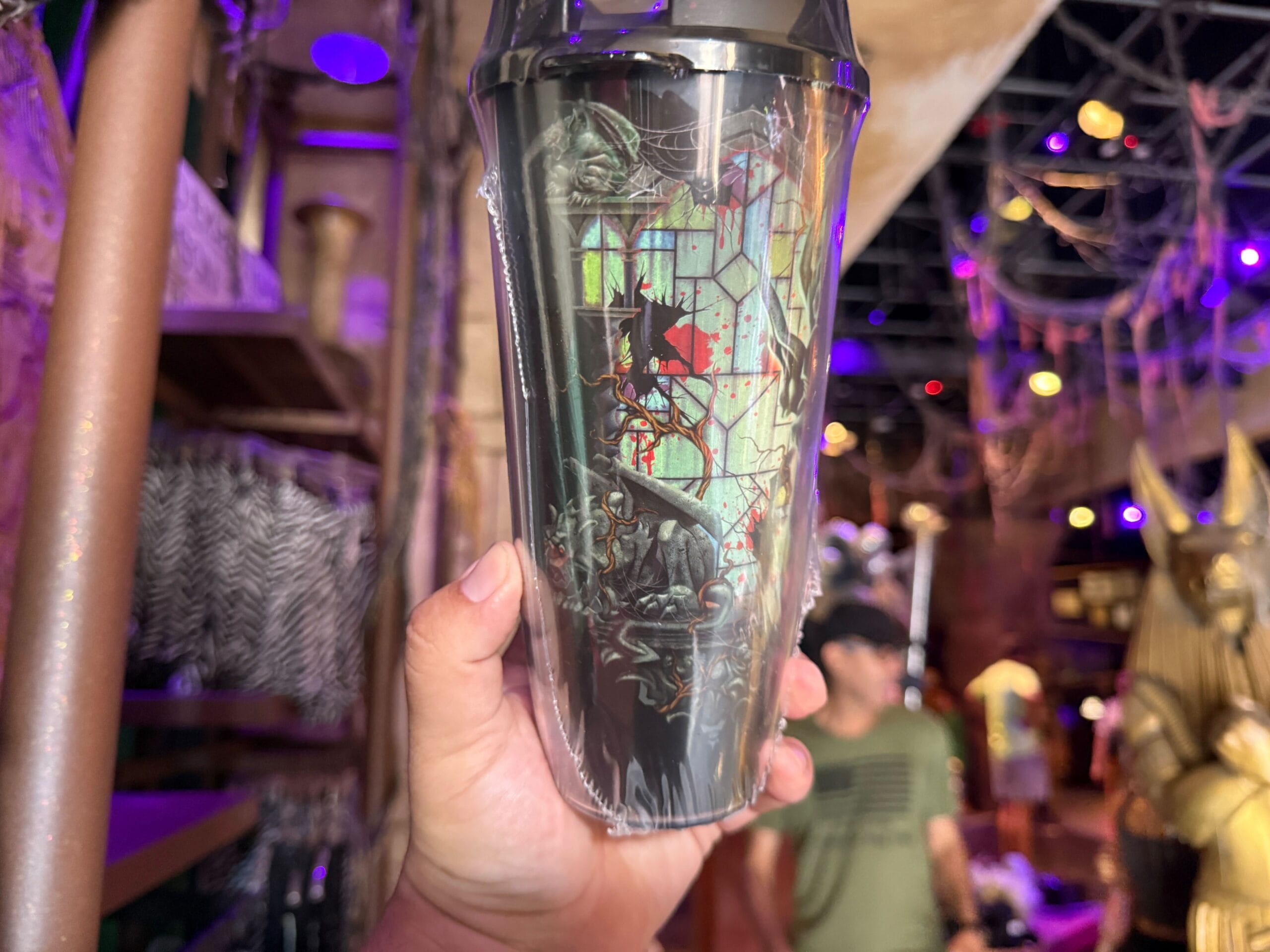A hand holds Halloween Horror Nights 34 merchandise—a dark tumbler with a red star, amid purple lighting and Egyptian decor.