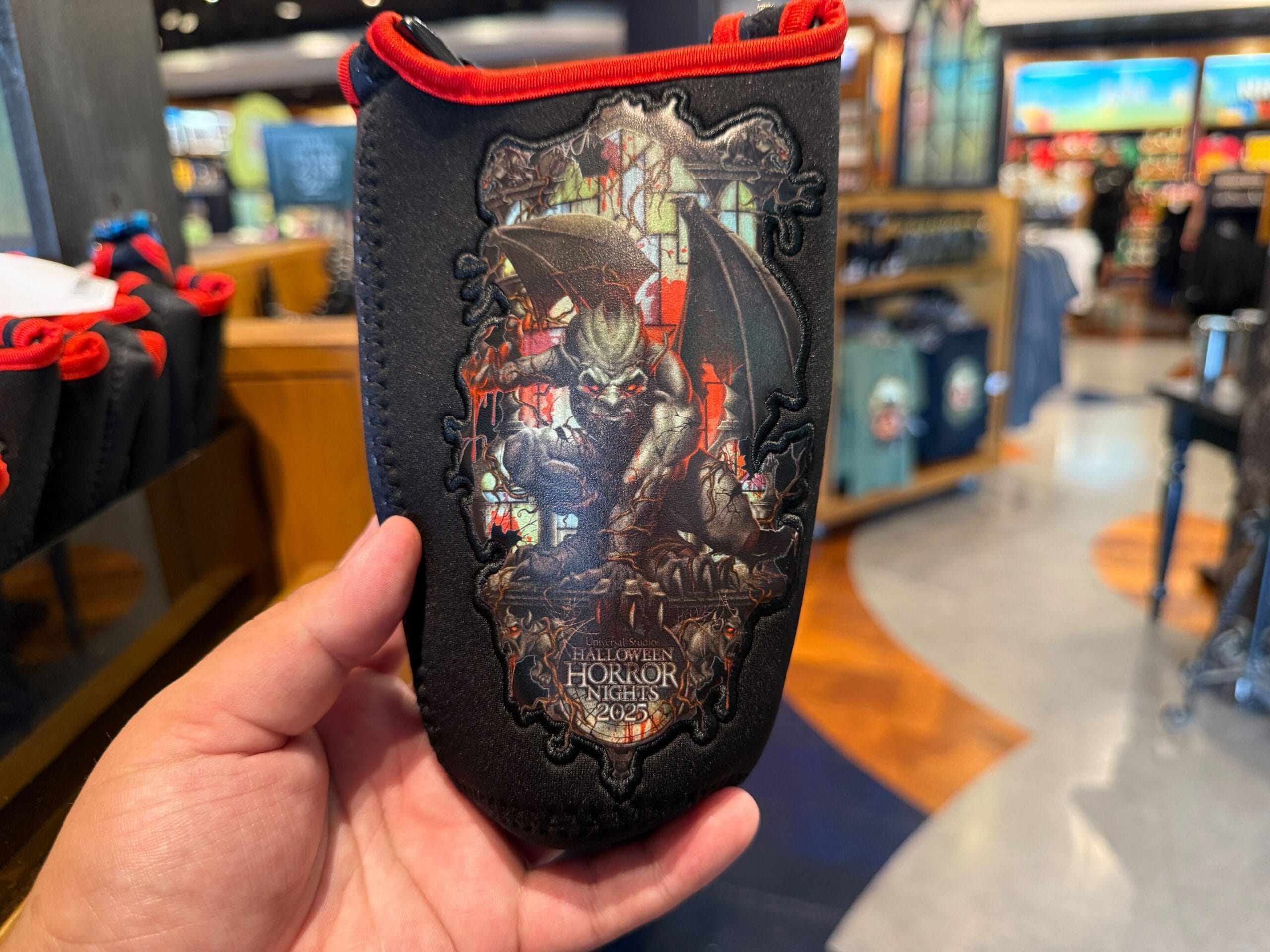 A hand holds HHN Merch: a black Halloween Horror Nights 2023 pouch with a menacing gargoyle and red accents.