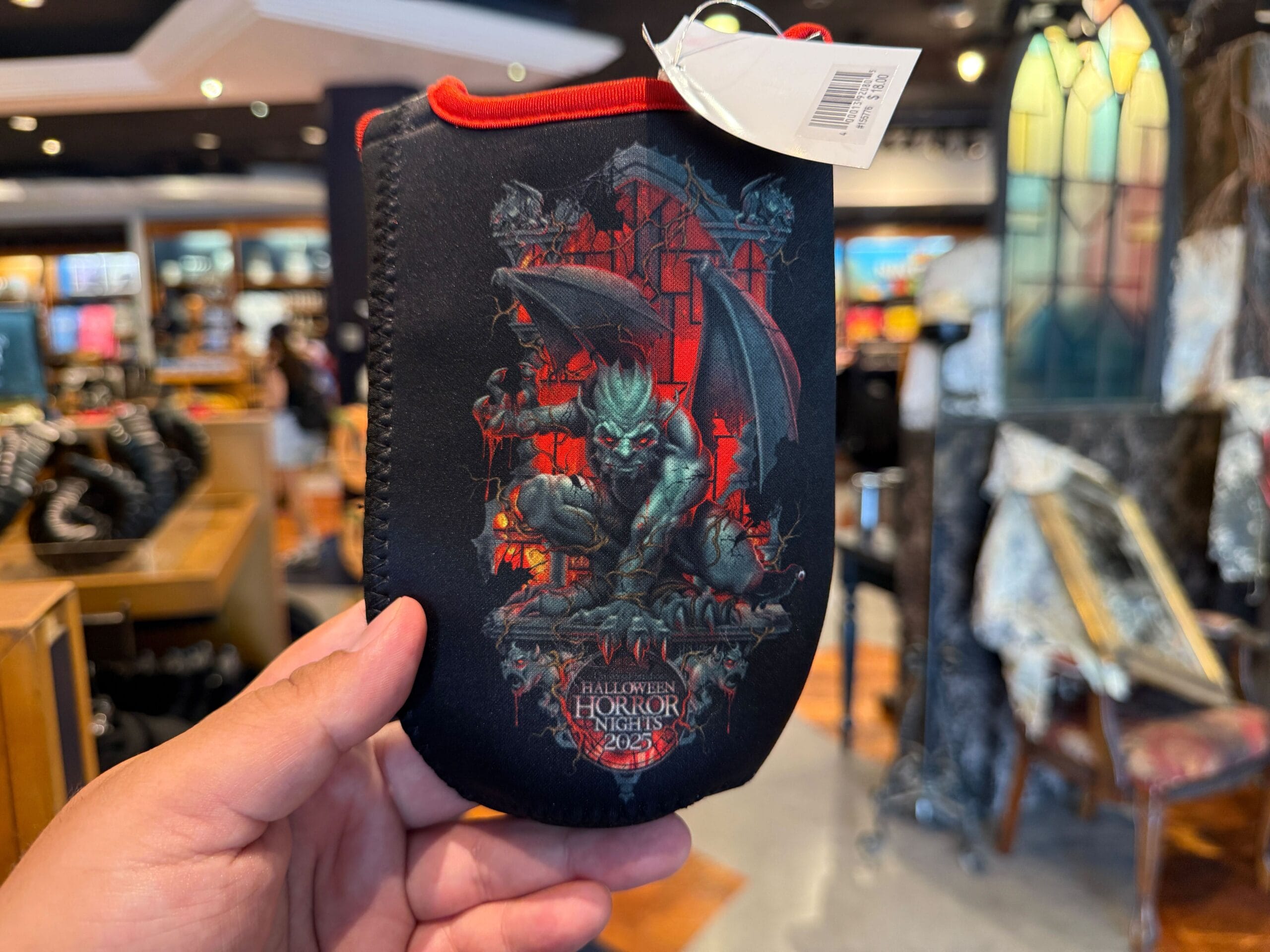 A hand holds an HHN Merch pouch with a fierce gargoyle illustration in a spooky Universal Orlando Resort store.