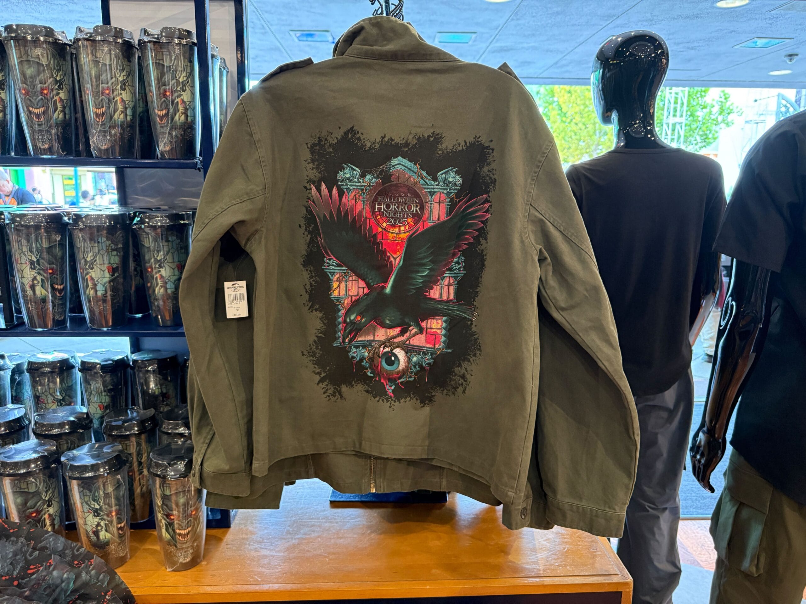 Olive jacket with a graphic of a raven and eye, and "Universal's Horror Hotel" text, displayed on a store table.