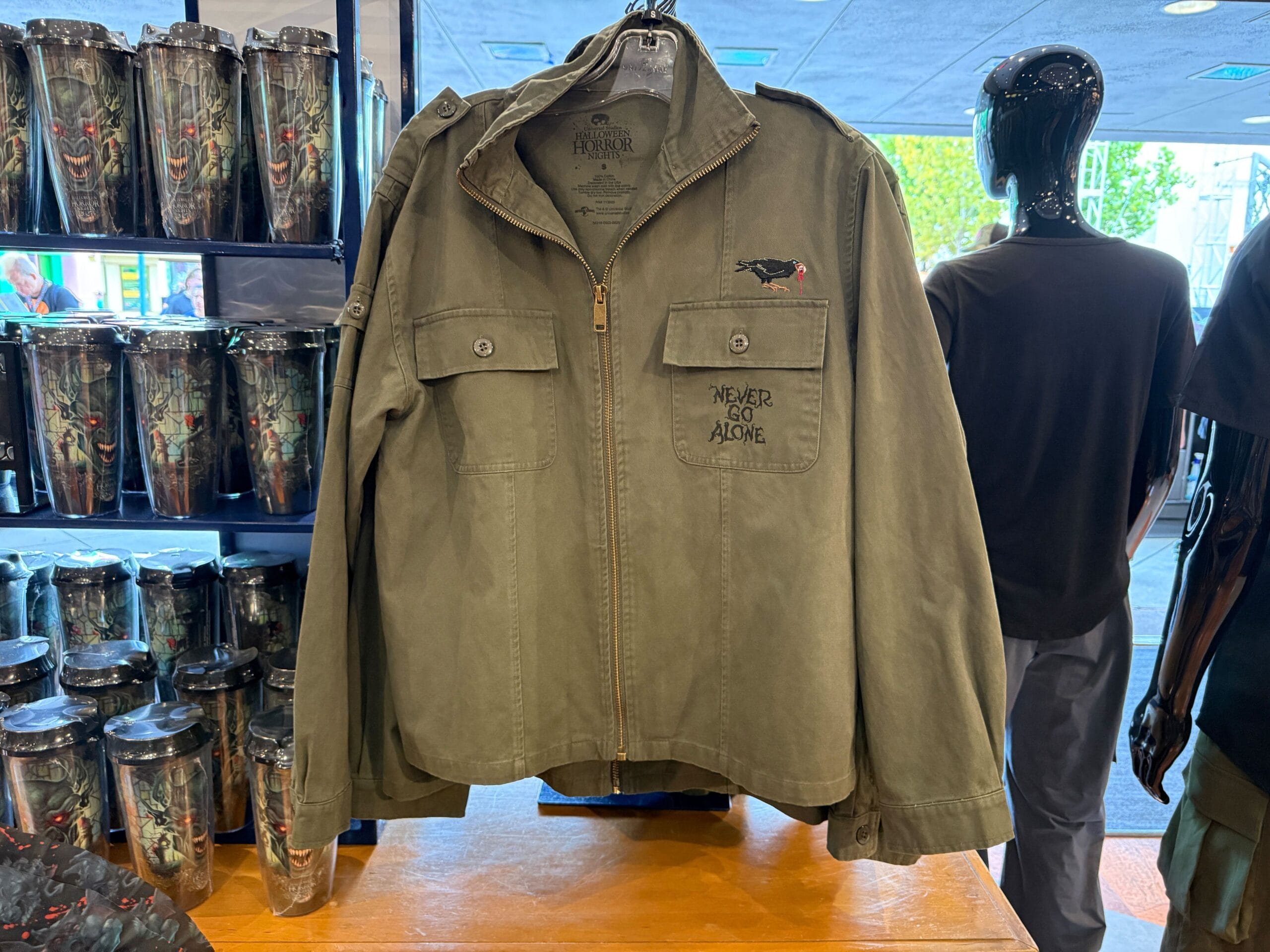 An olive green jacket with "NEVER GO ALONE" text is displayed on a rack in a store next to cups and shirts.