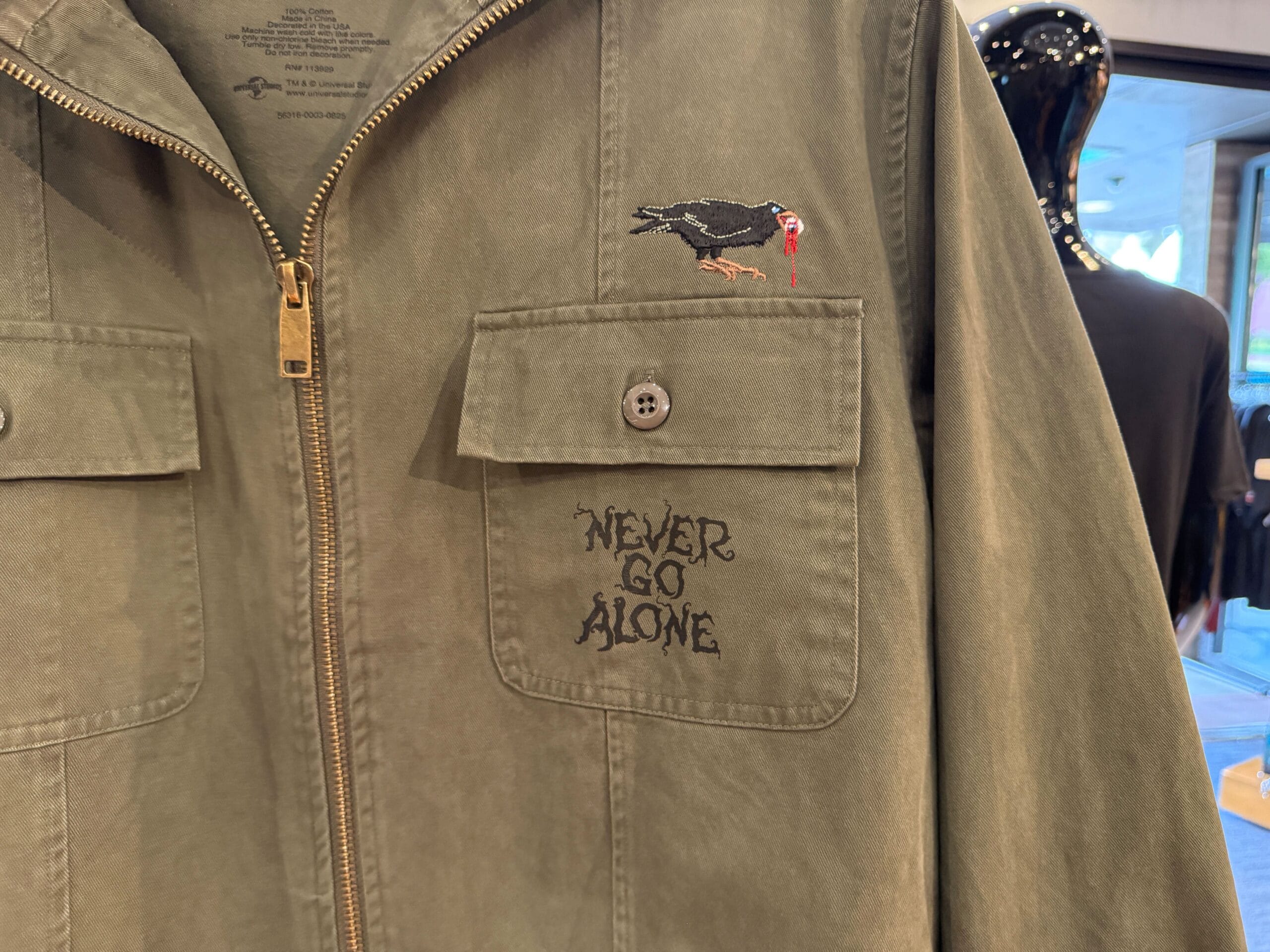 hhn merch 2025 Olive jacket with "NEVER GO ALONE" text and an embroidered bird with a red beak above the left pocket.