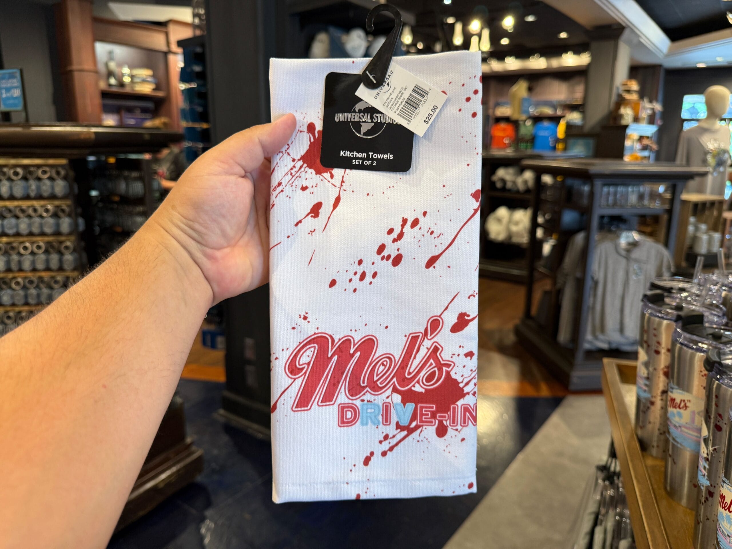 A hand holds a Mel's Drive-In kitchen towel with red splatter design in a store.