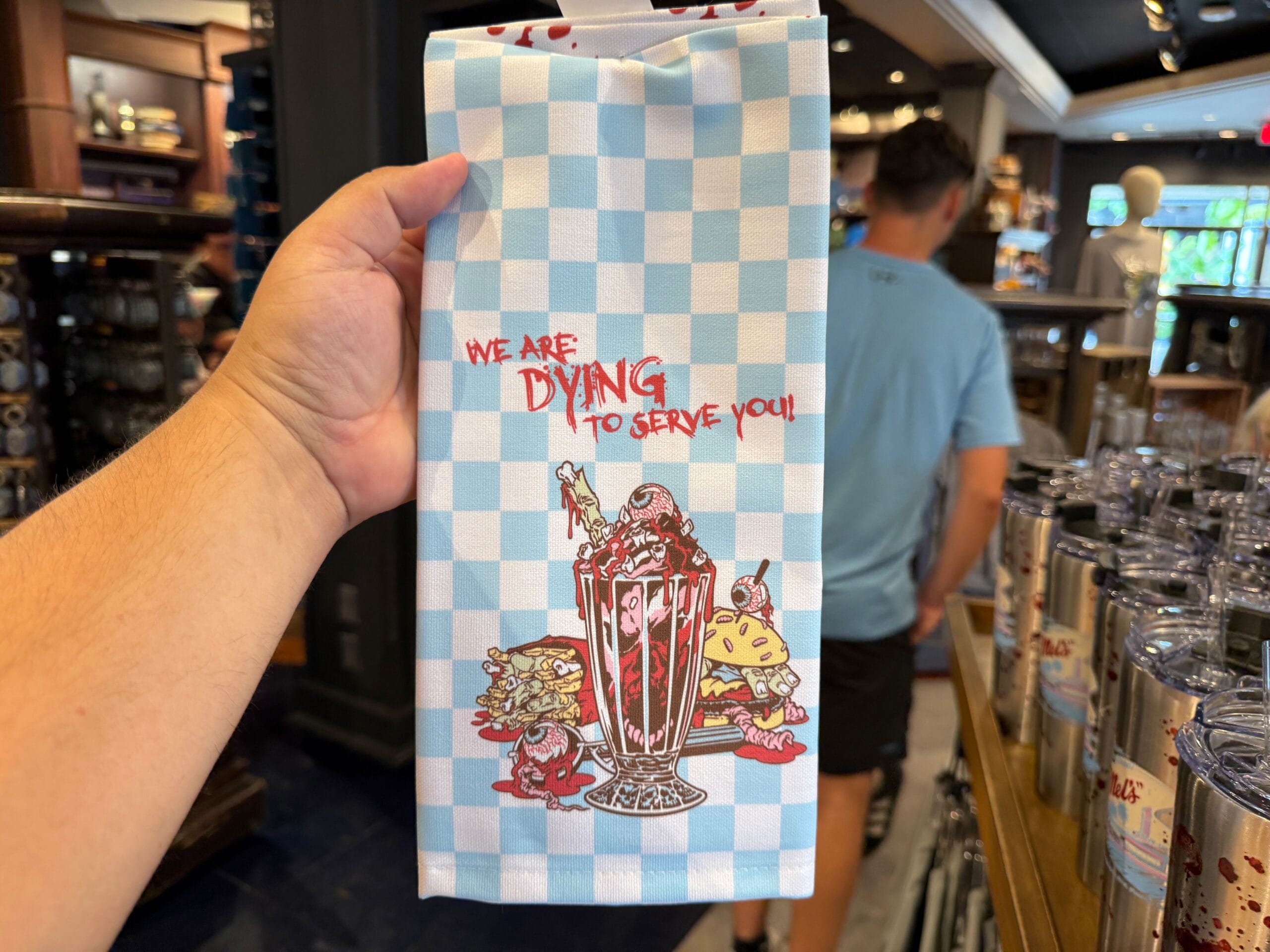 A hand holds a towel with a milkshake and food cartoon, reading "We are dying to serve you!" on blue-checkered fabric.