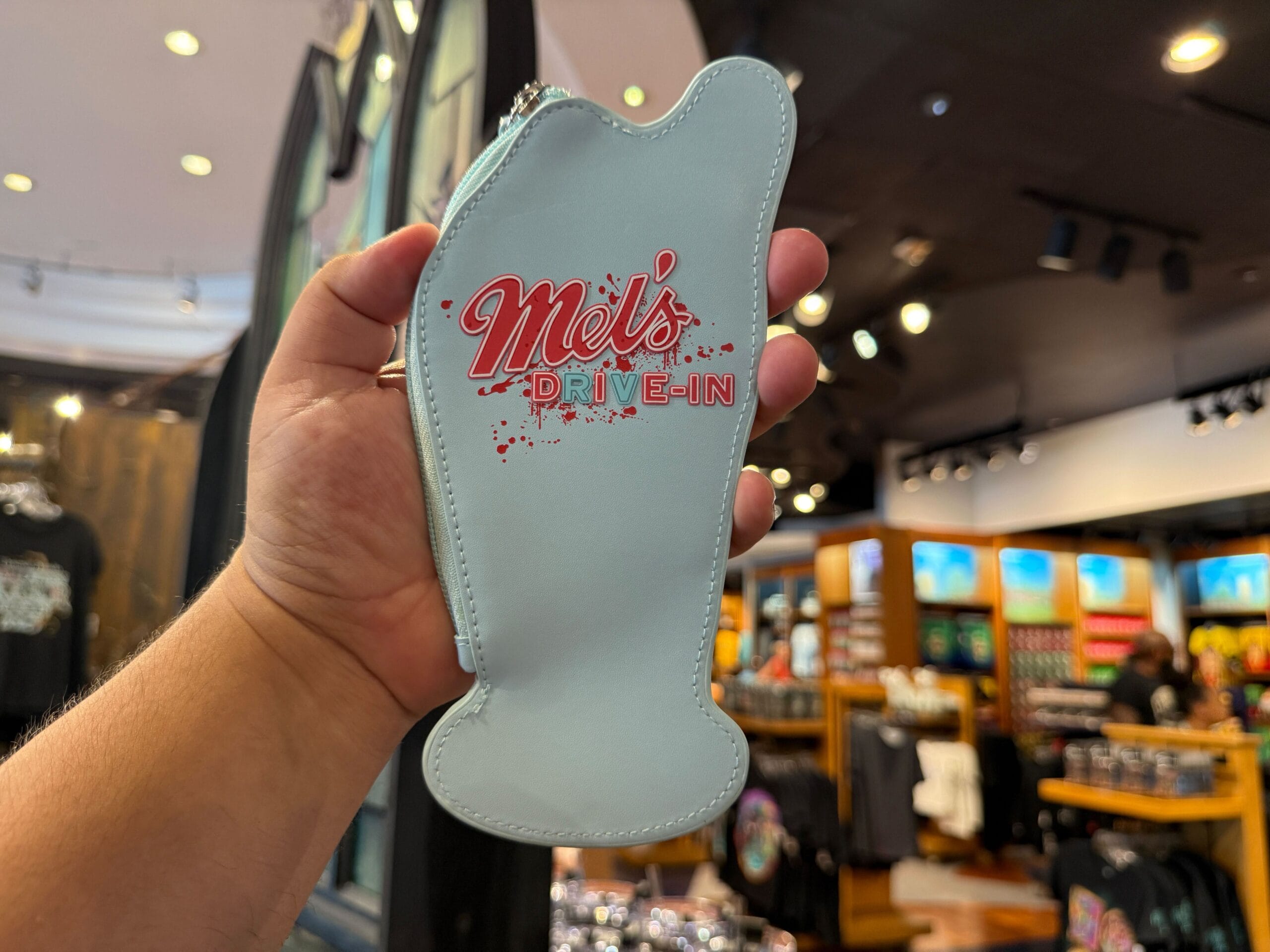 A hand holds a milkshake-shaped coin purse that says "Mel's Drive-In" inside a store with shirts on display.