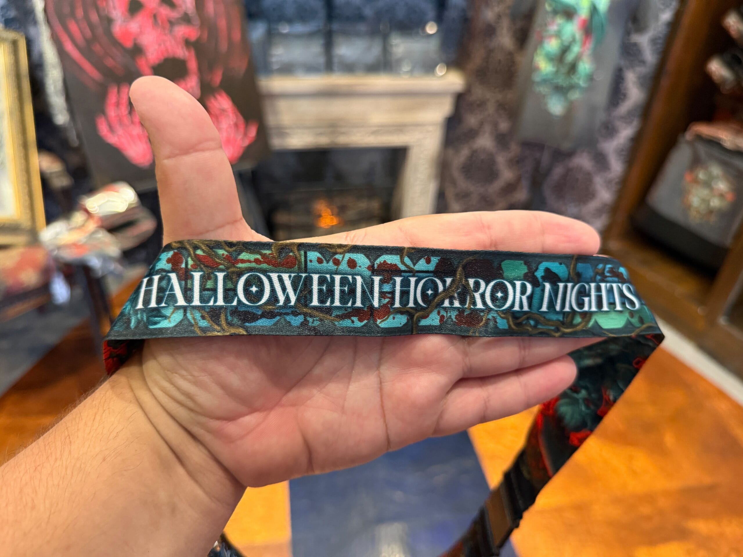 A hand holds an HHN merch lanyard in a decorated room at Universal Orlando Resort with a fireplace in the background.