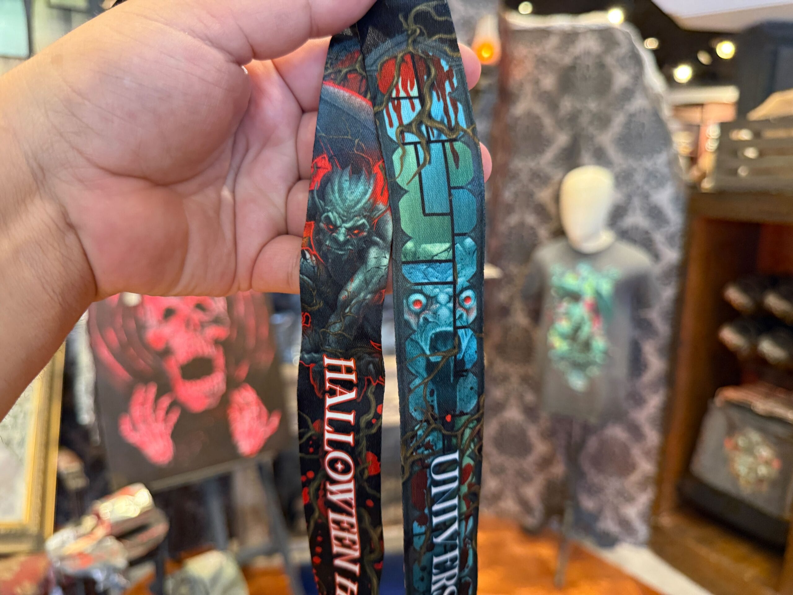 A hand holds a Halloween Horror Nights 2025 lanyard with horror designs in a store selling spooky merch at Universal Orlando Resort.
