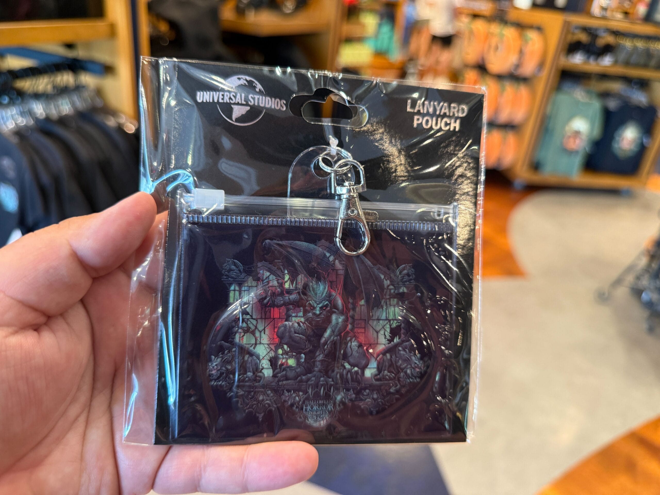 A hand holds a packaged Halloween Horror Nights 2025 lanyard pouch with a dark, gothic design in a Universal Orlando Resort store.
